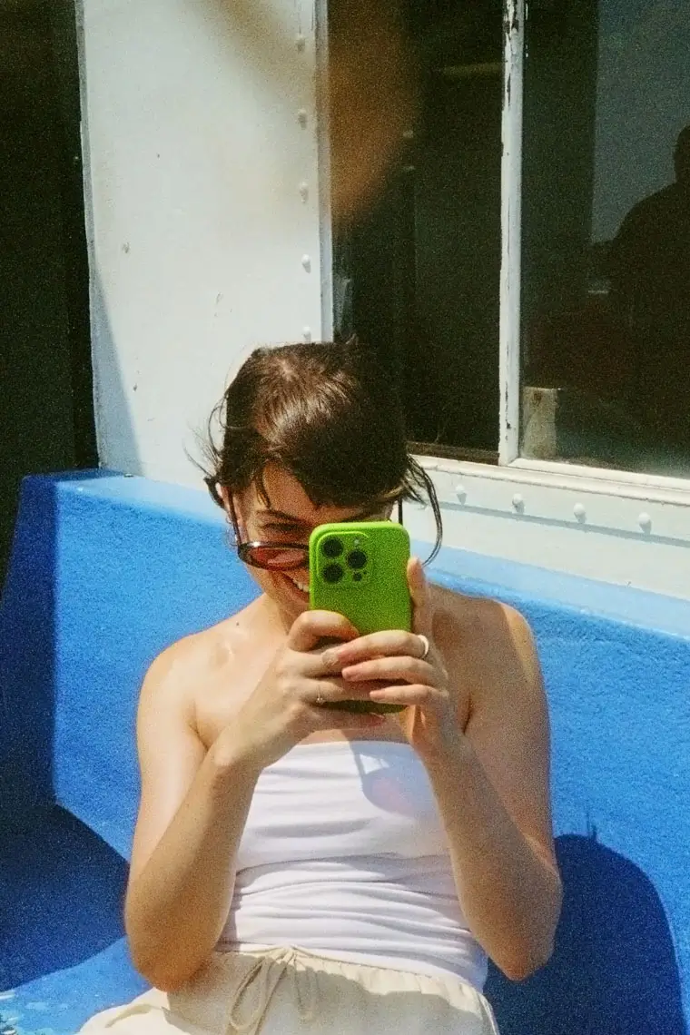 Young woman sitting on a blue bench, smiling while taking a photo with her green smartphone.