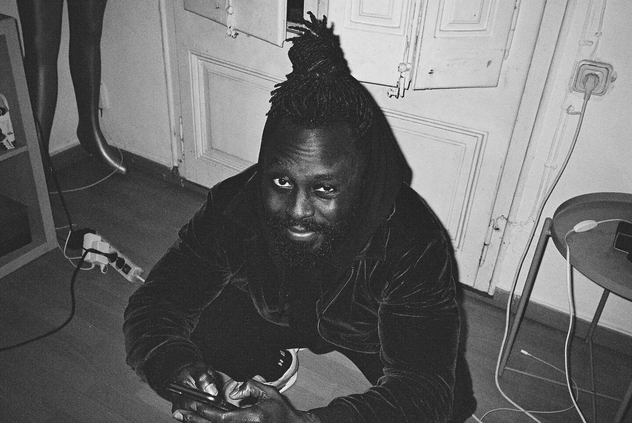Black and white photo of a man with dreadlocks squatting and smiling while holding a phone indoors.