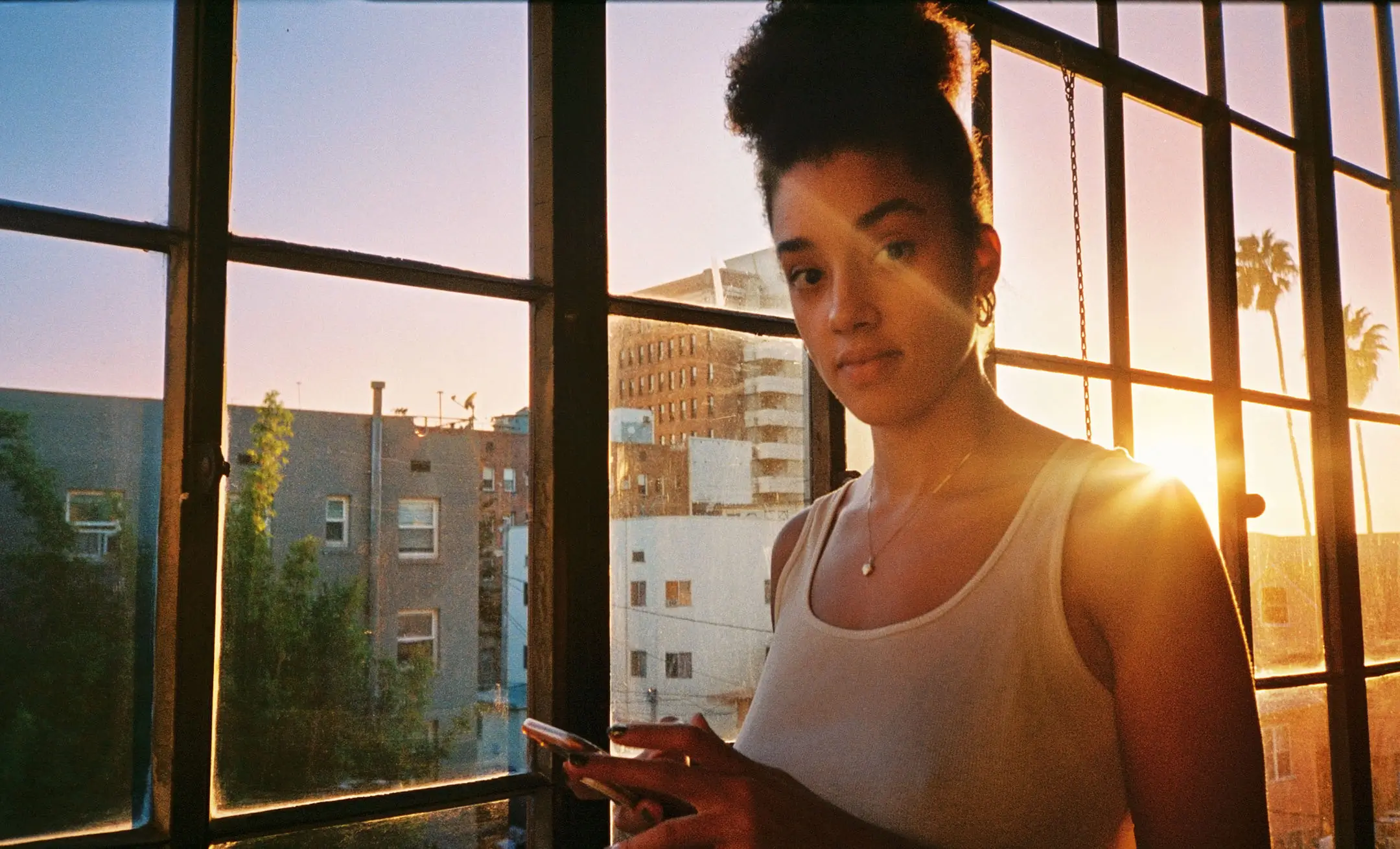 Young woman with curly hair in a bun using a smartphone by a large window at sunset with city buildings and palm trees outside.
