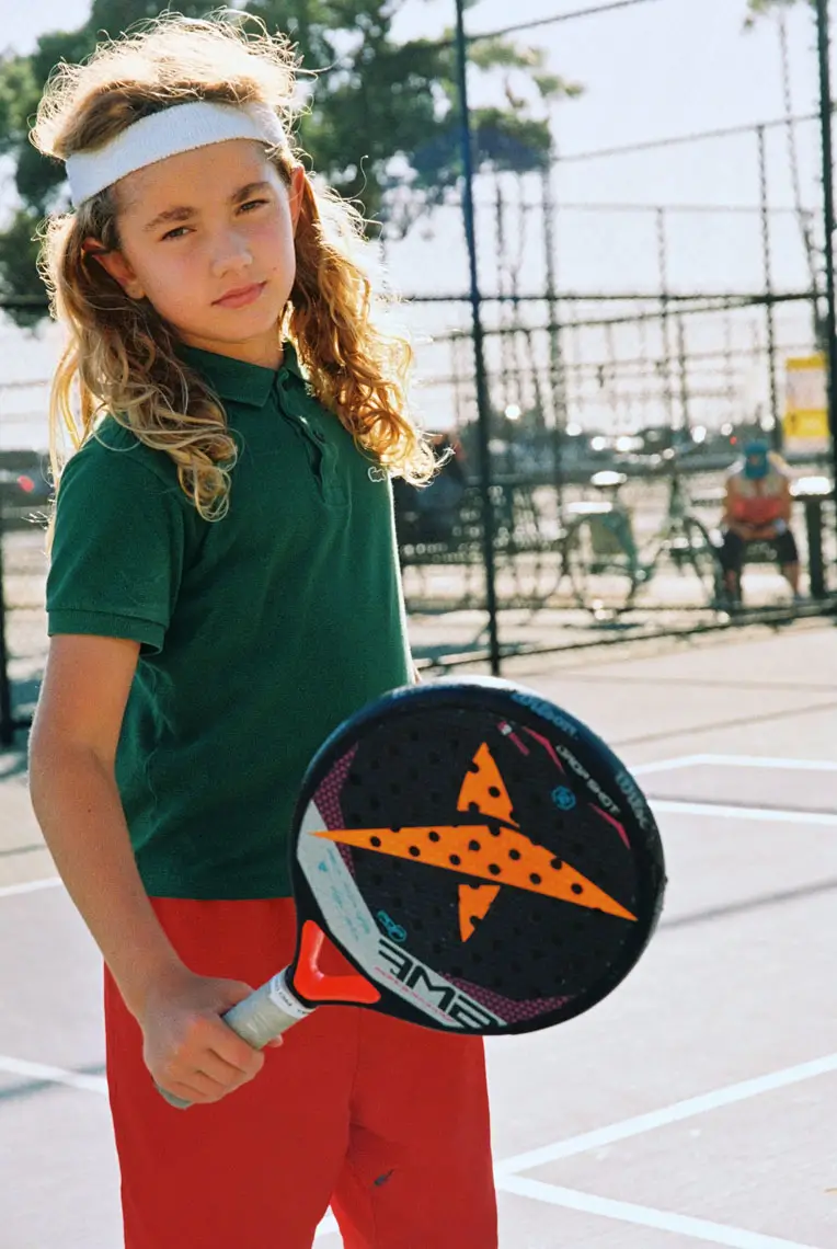 Young player with long curly hair wearing a headband holding a paddle on an outdoor padel court.