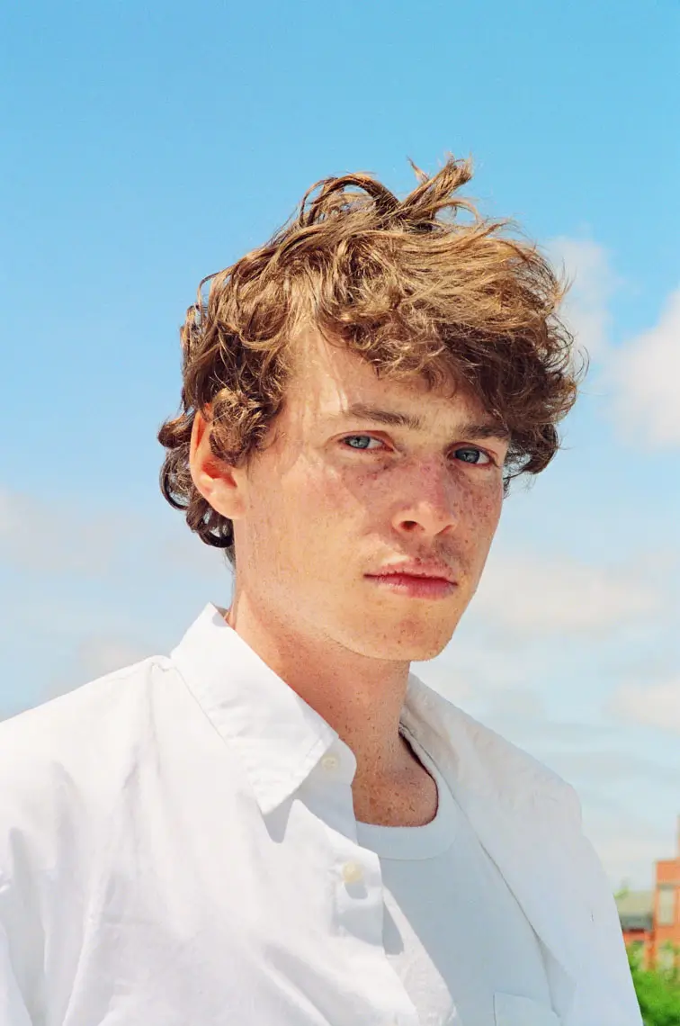 Portrait of a young man with curly brown hair and freckles, wearing a white shirt against a blue sky background.