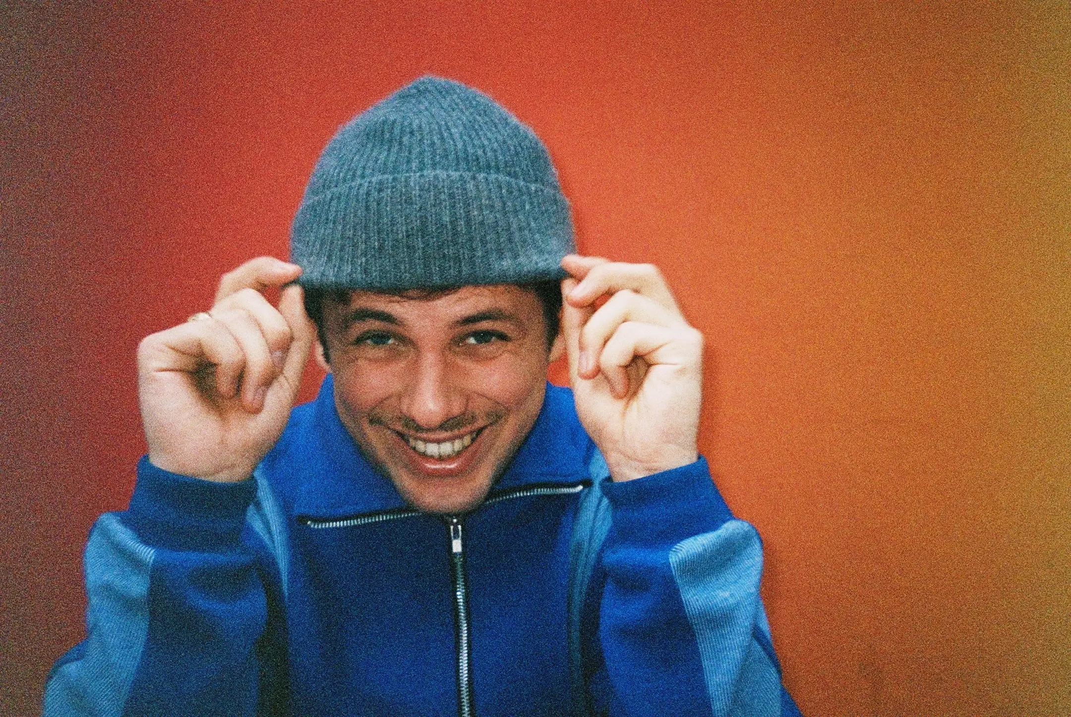 Smiling man wearing a blue jacket and a gray knit beanie against a gradient red-orange background.