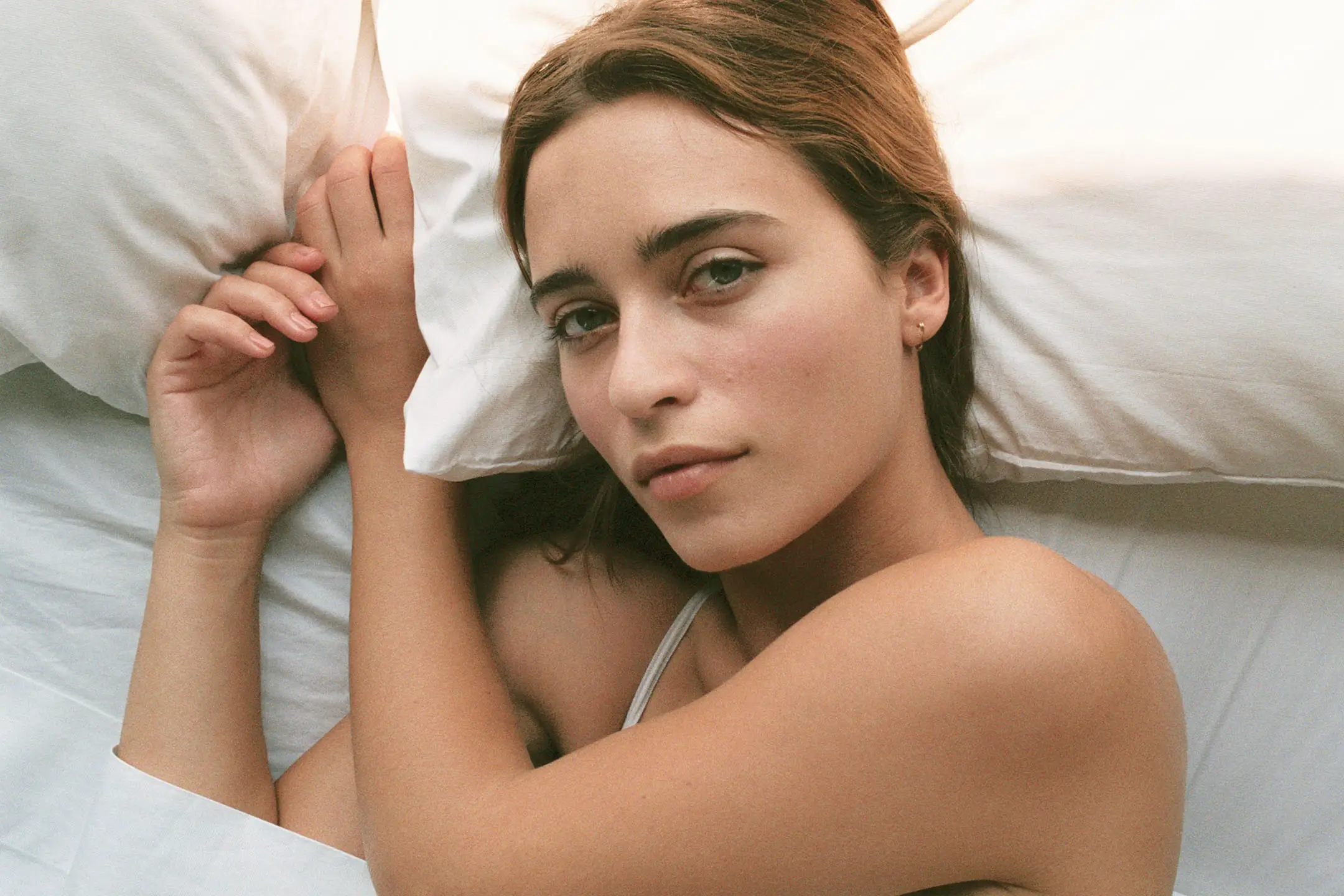 Young woman with brown hair and green eyes resting on white bed sheets, looking calmly at the camera.