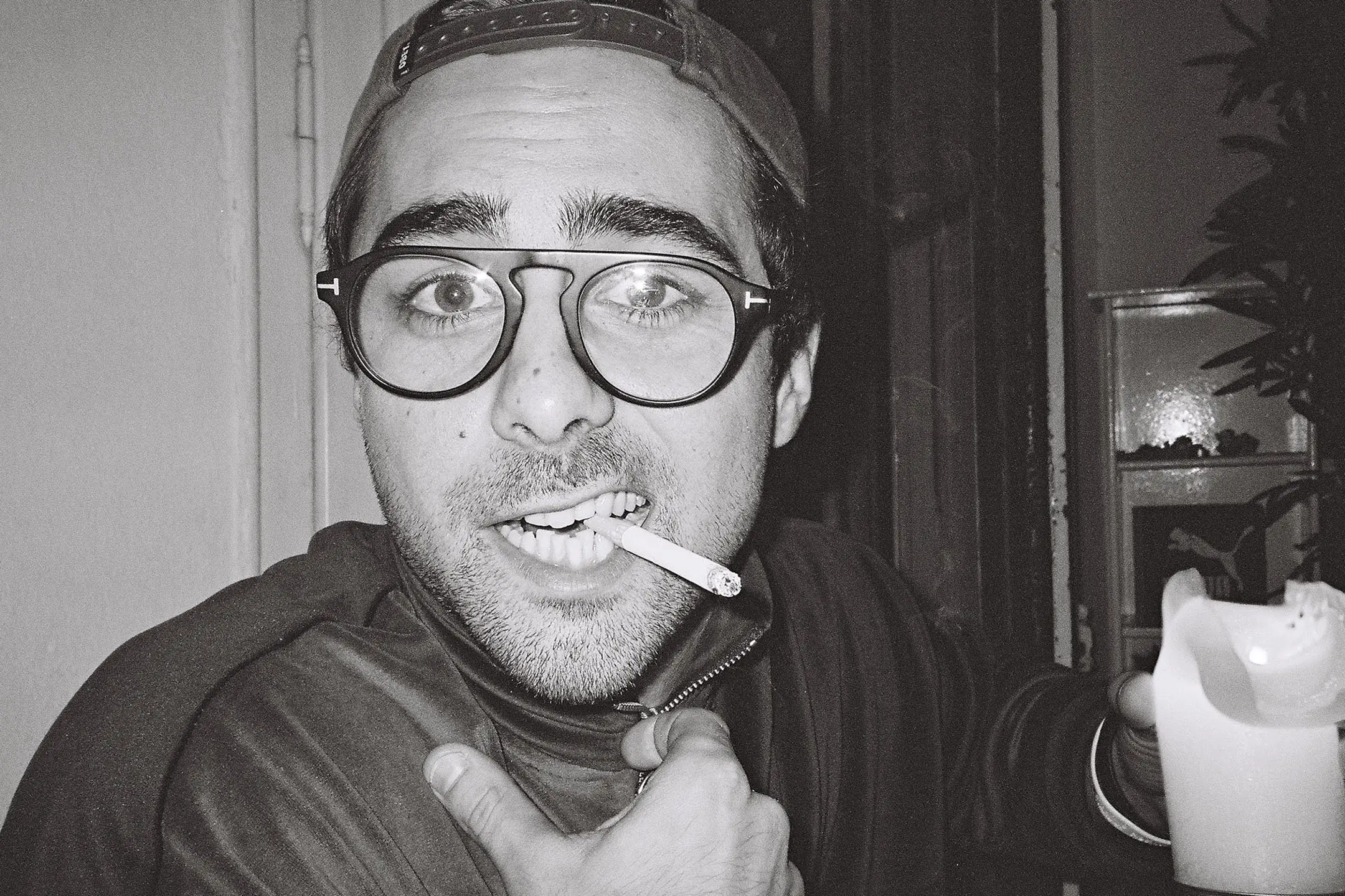 Man wearing glasses and a backwards cap with a cigarette in his mouth, holding up a white cup and looking at the camera.