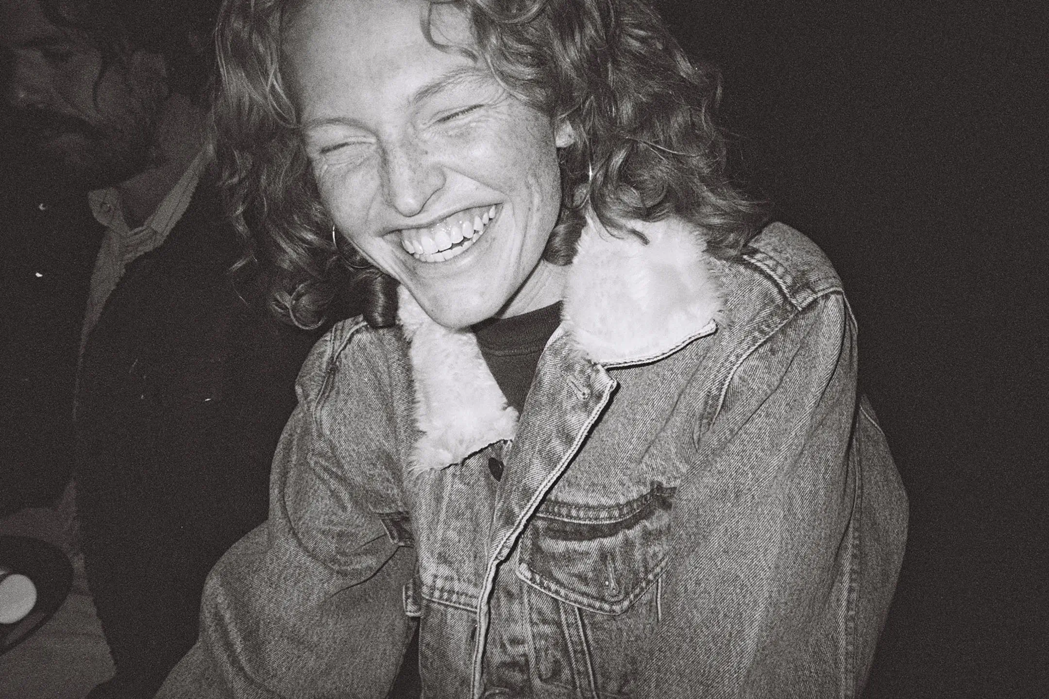 Close-up of a woman with curly hair laughing with eyes closed, wearing a denim jacket with a fluffy collar.
