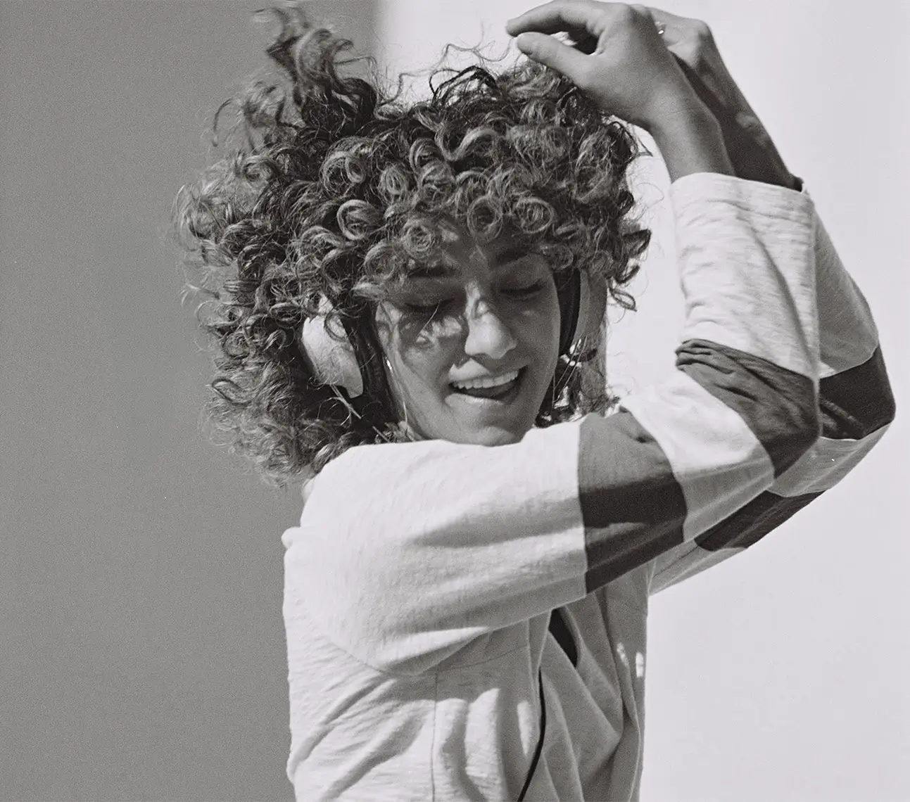 Person with curly hair wearing headphones and a long-sleeve shirt with dark stripes, smiling with eyes closed.