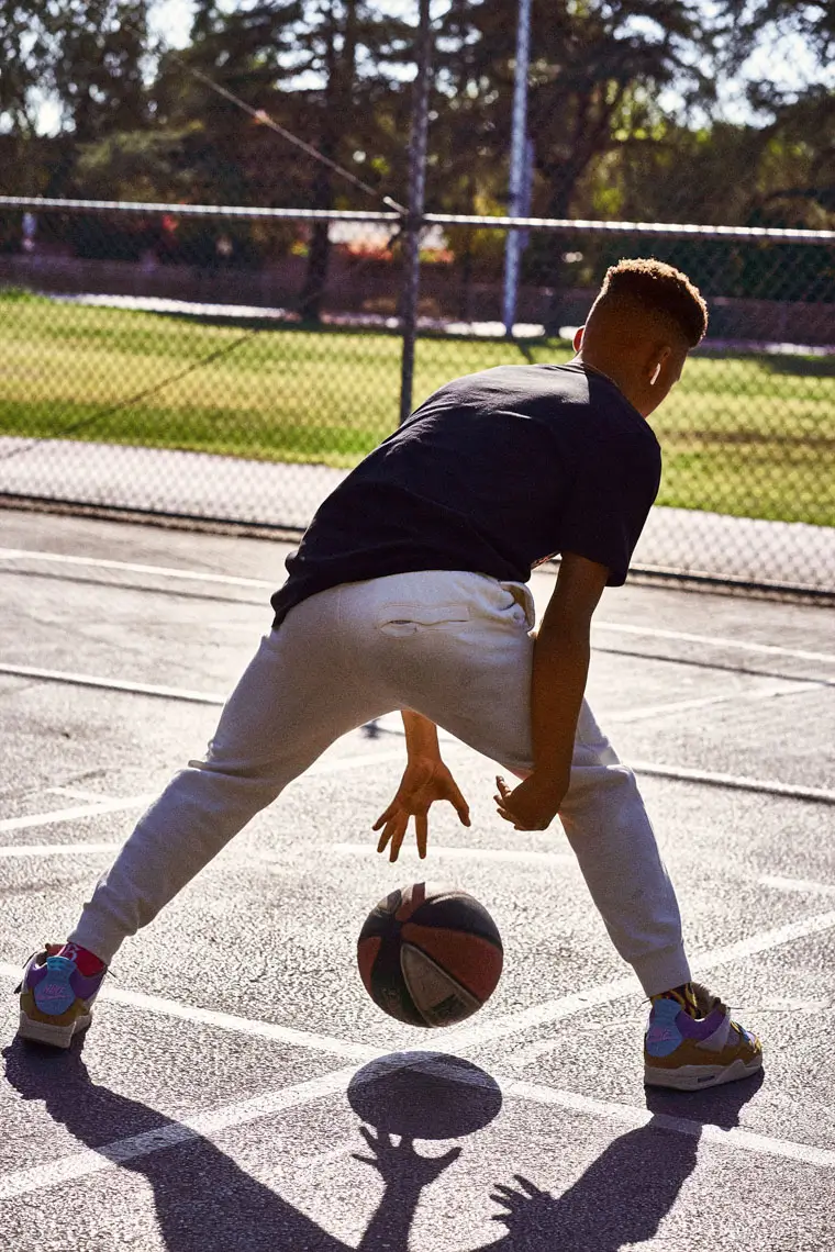 Young man dribbling a basketball low on an outdoor court in front of a chain-link fence.