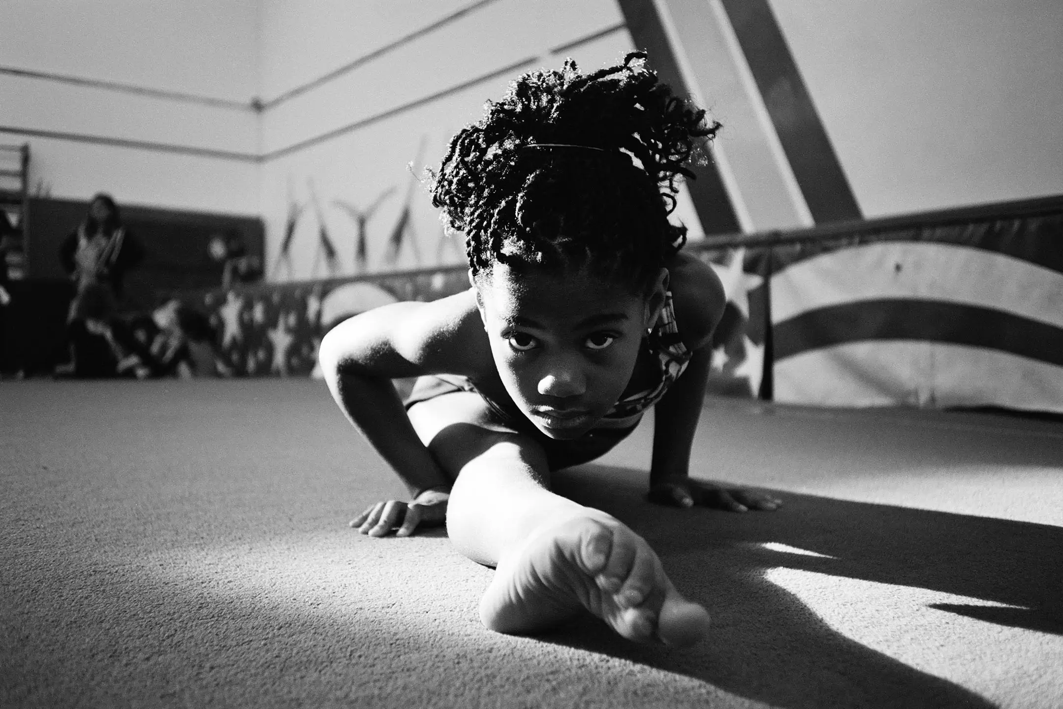 Young female gymnast with braided hair in a gym performing a stretch on the floor, looking directly at the camera.