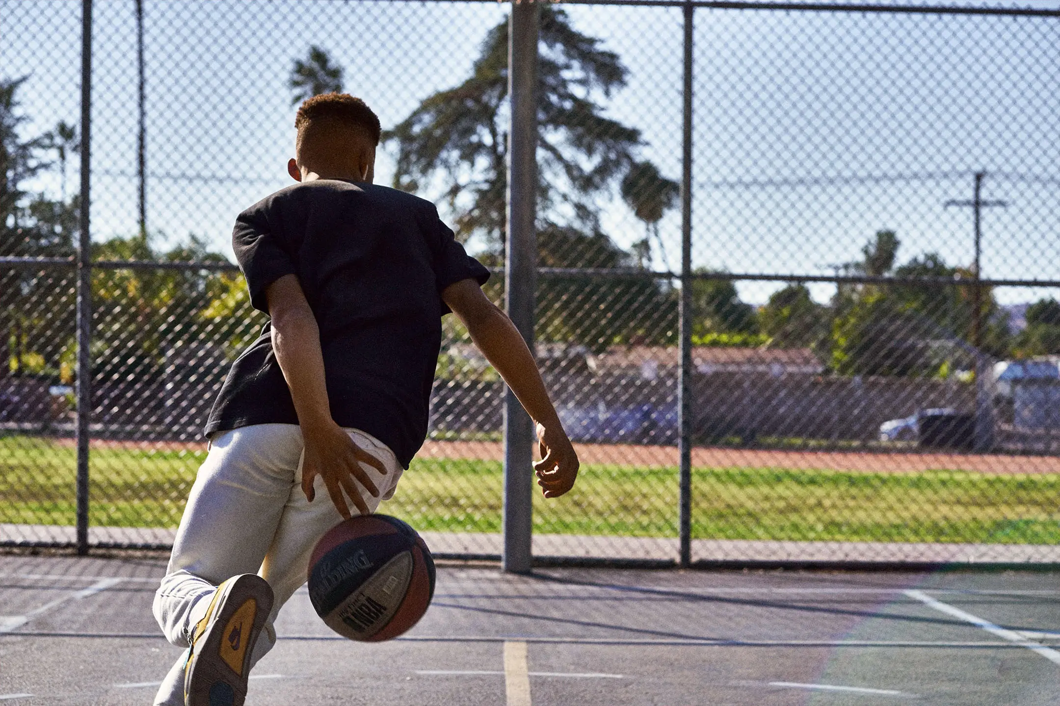 Young basketball player dribbling a basketball on an outdoor court with a chain-link fence and trees in the background.