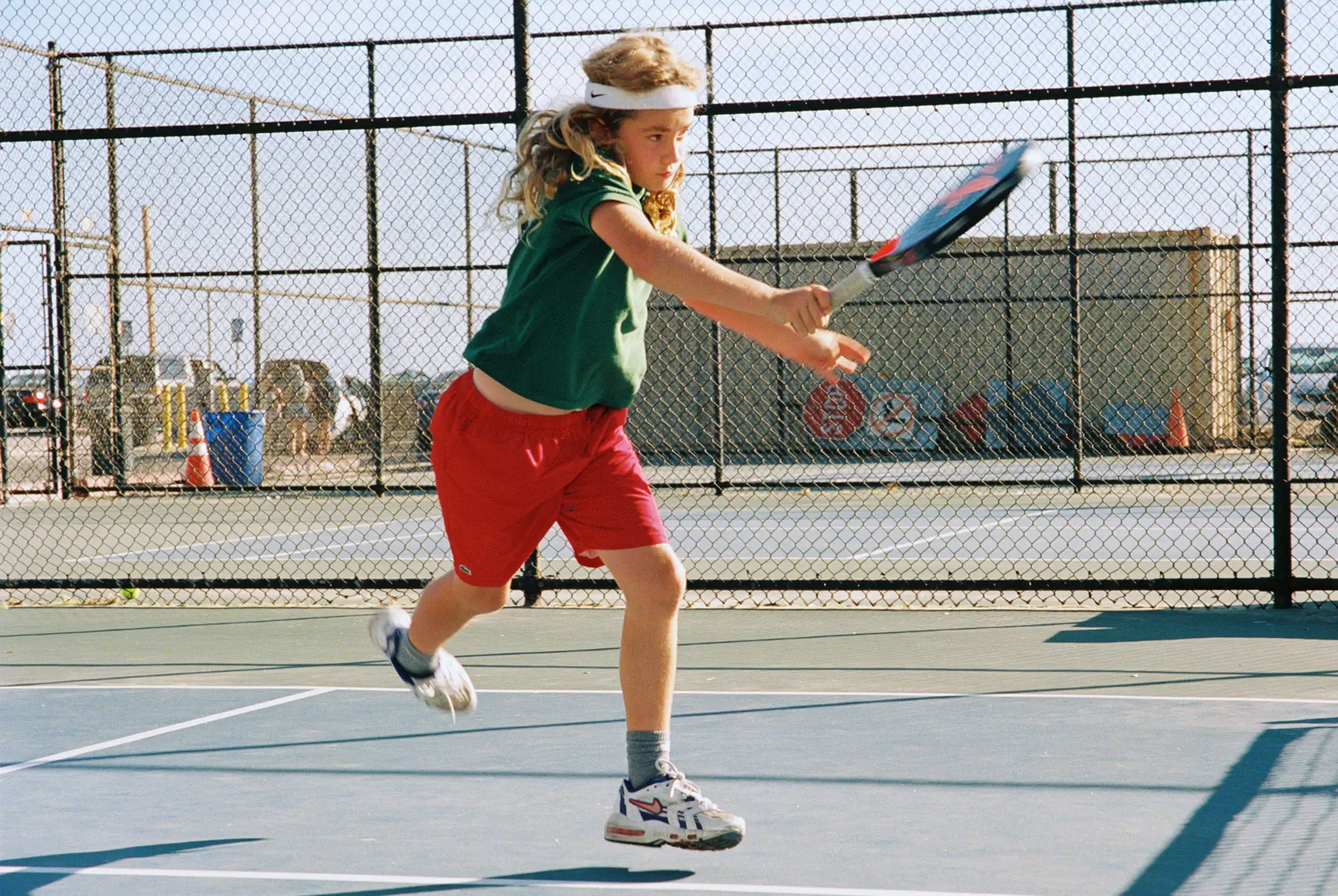 Young girl in red shorts and green shirt hitting a tennis ball on an outdoor court with a chain-link fence background.