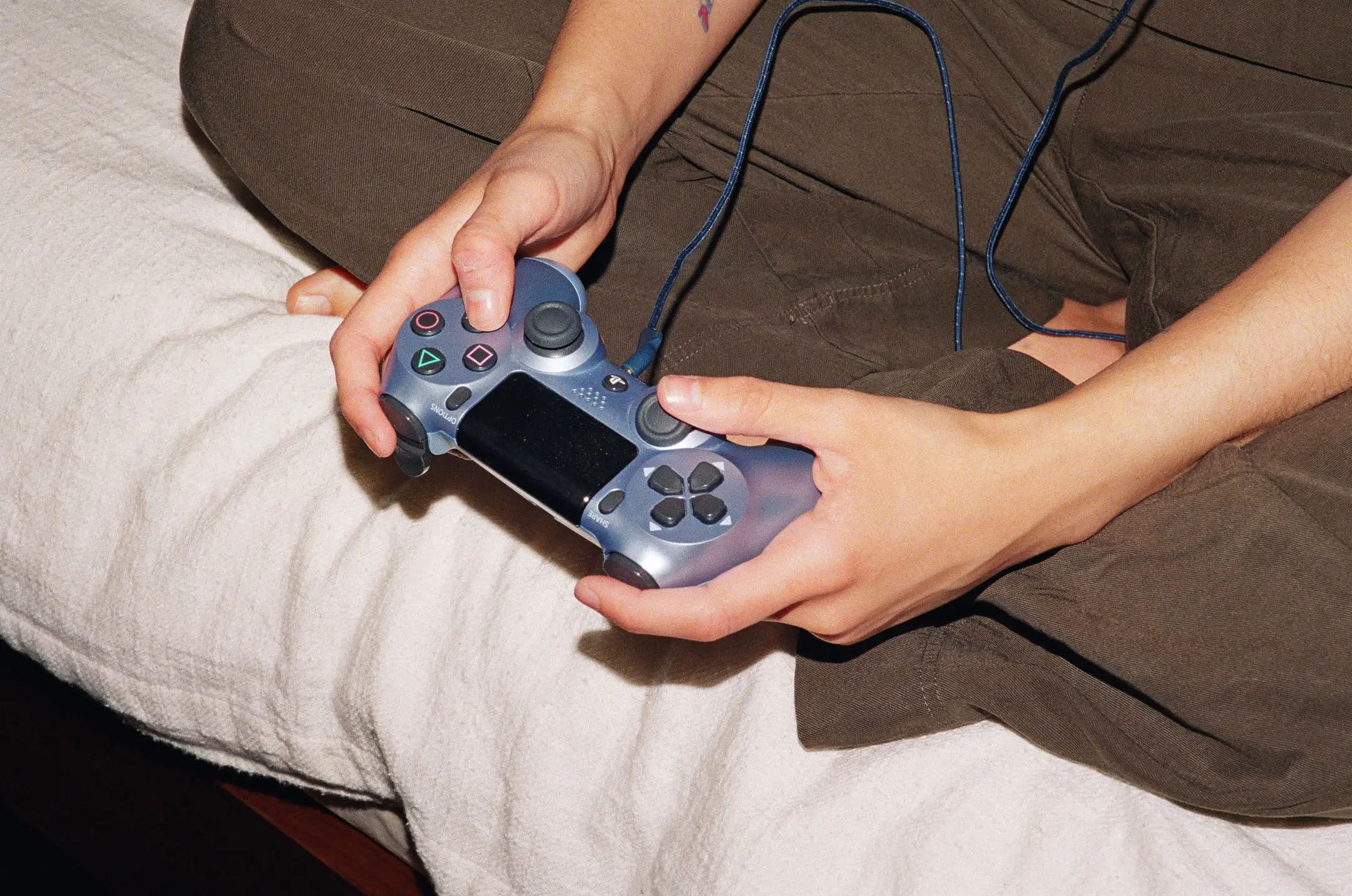 Close-up of hands holding a wired PlayStation controller while sitting cross-legged on a bed.