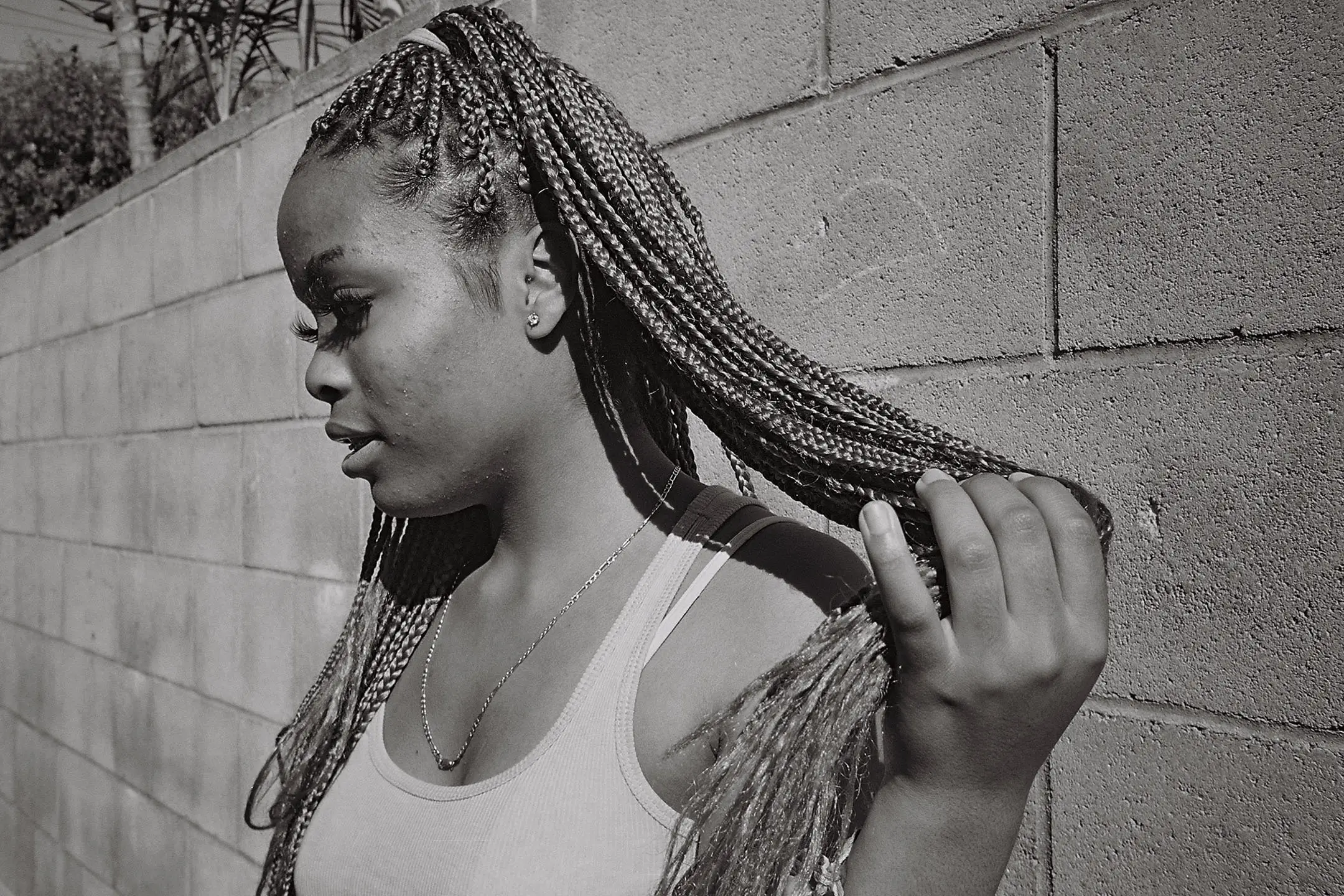 Side profile of a woman with long braided hair holding the ends against a stone wall background.