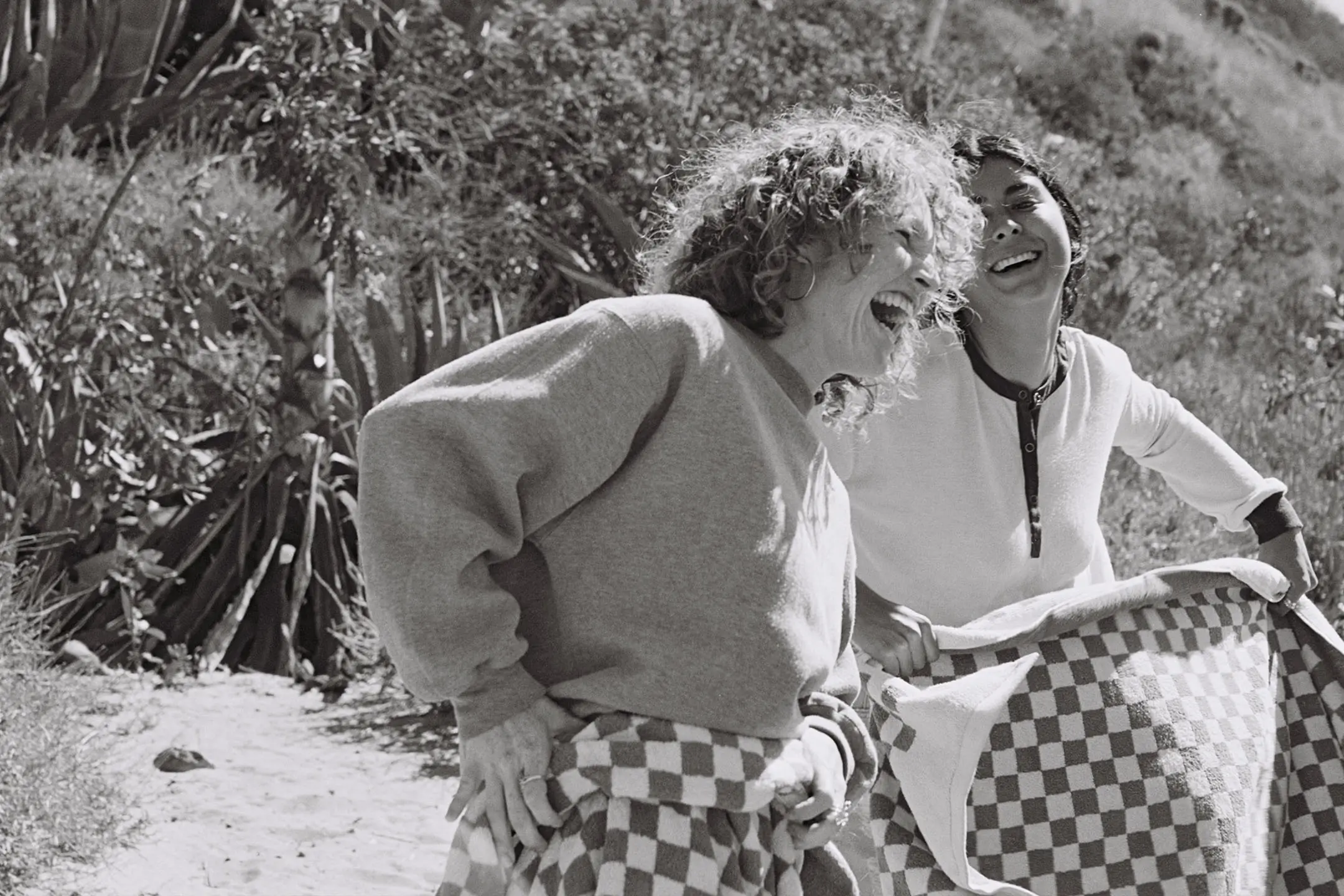 Two women laughing and holding a checkered blanket outdoors on a sunny day.