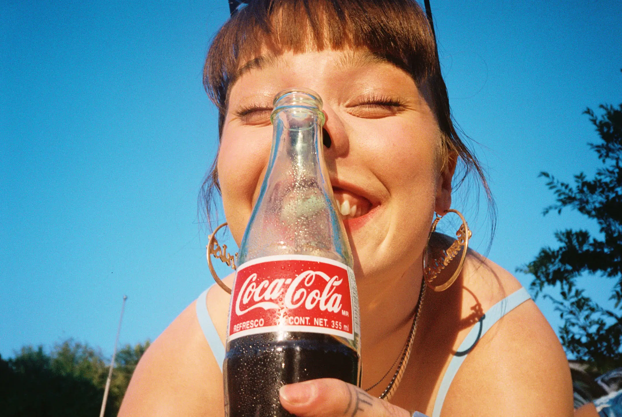 Smiling young woman holding a cold Coca-Cola glass bottle close to her face against a clear blue sky.