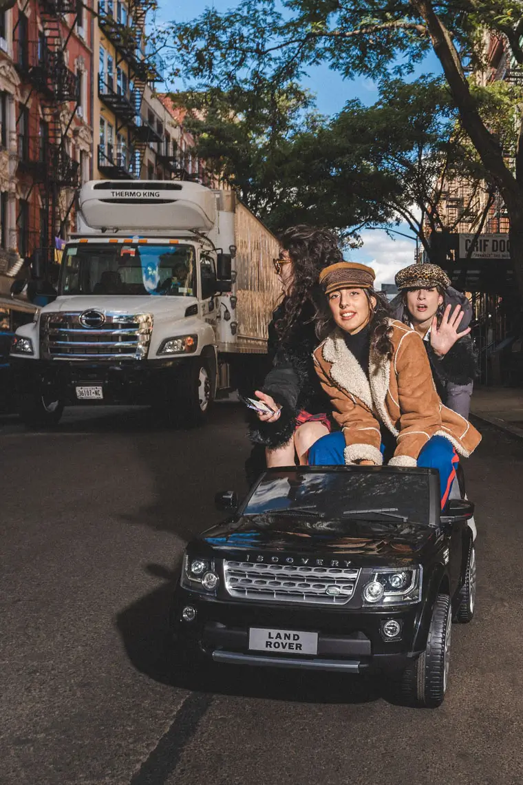 Three girls riding a miniature black Land Rover toy car on a city street with a white delivery truck and buildings in the background.