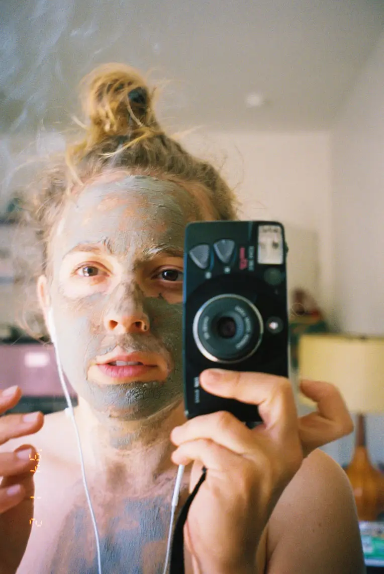 Person with a clay face mask taking a selfie in a mirror holding a camera, with hair tied up and earphones in.