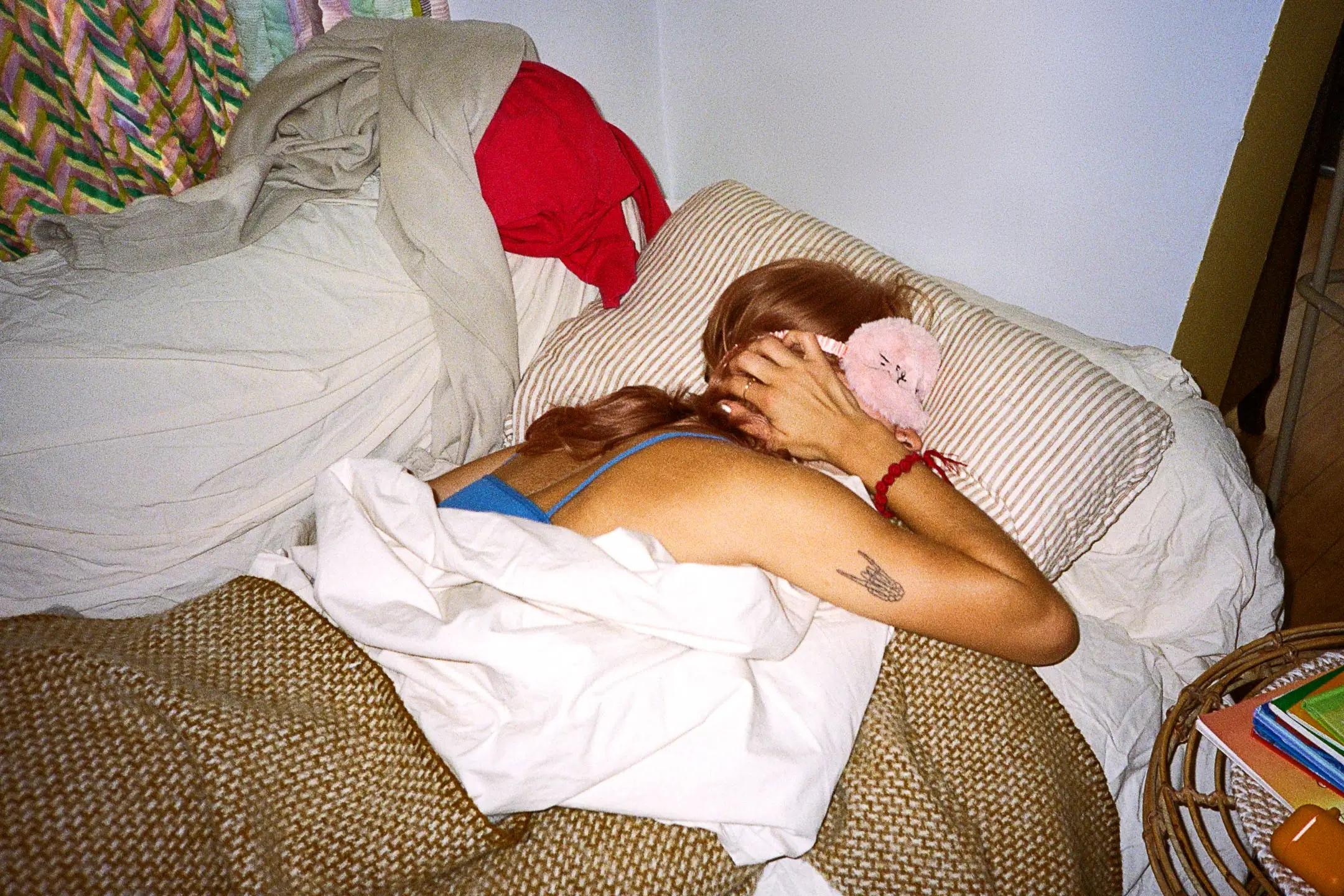 Person lying on a bed with a striped pillow, wearing a pink sleep mask and a blue top, covered with a brown and white blanket.