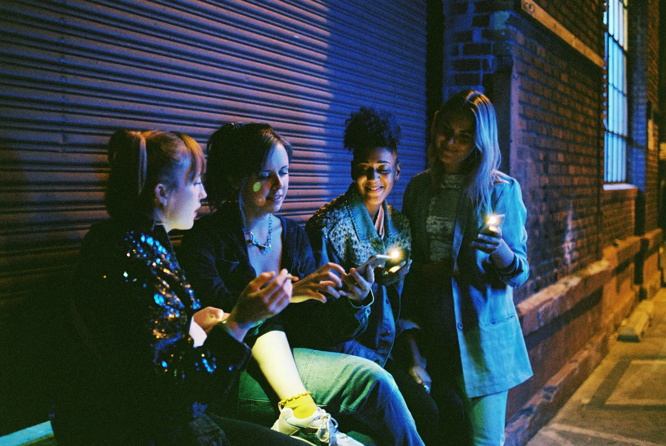 Four young women sitting and standing near a brick wall at night, illuminated by their phone and lighter lights.