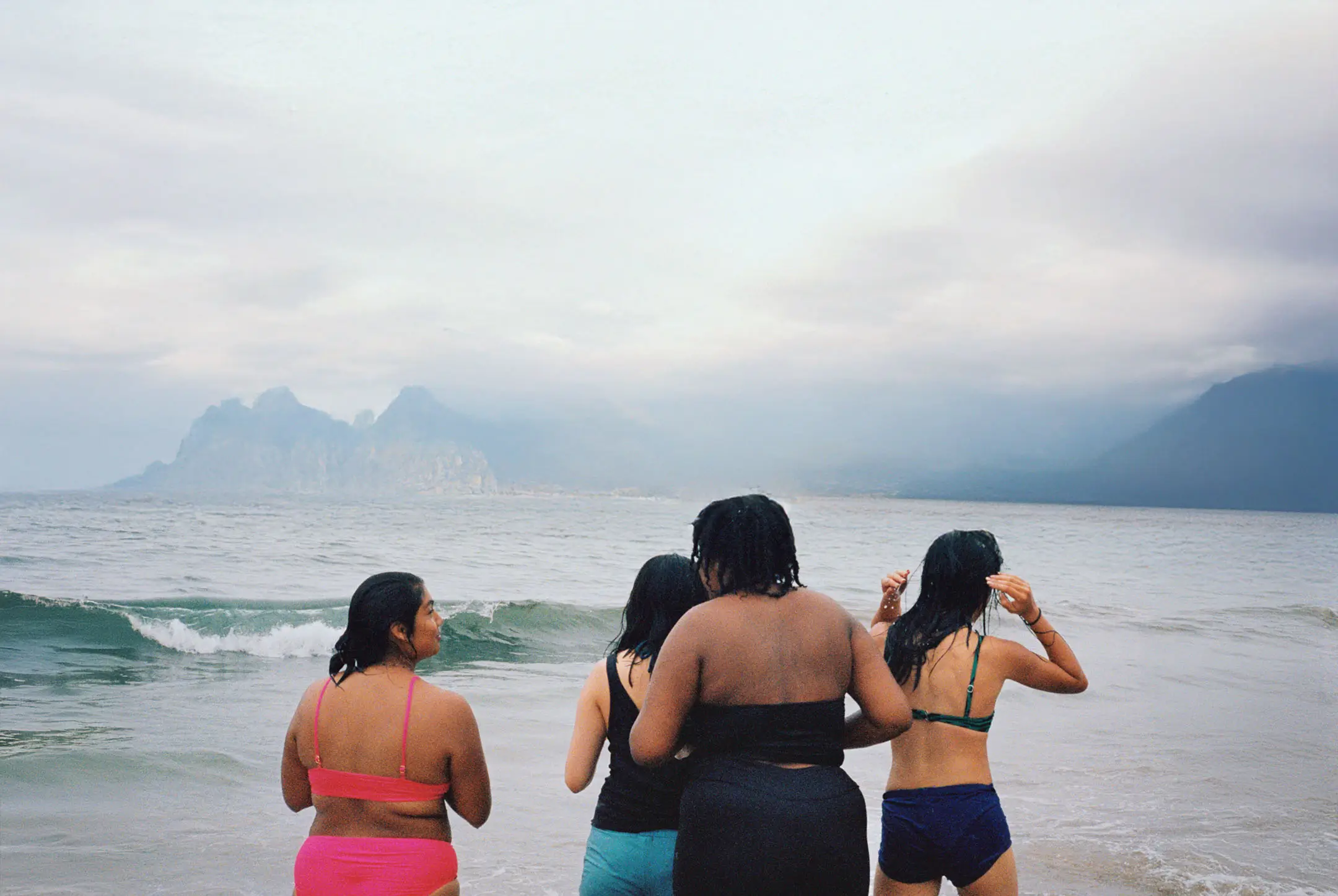 Four women in swimsuits standing at the edge of the ocean with mountainous coastline in the background under a cloudy sky.