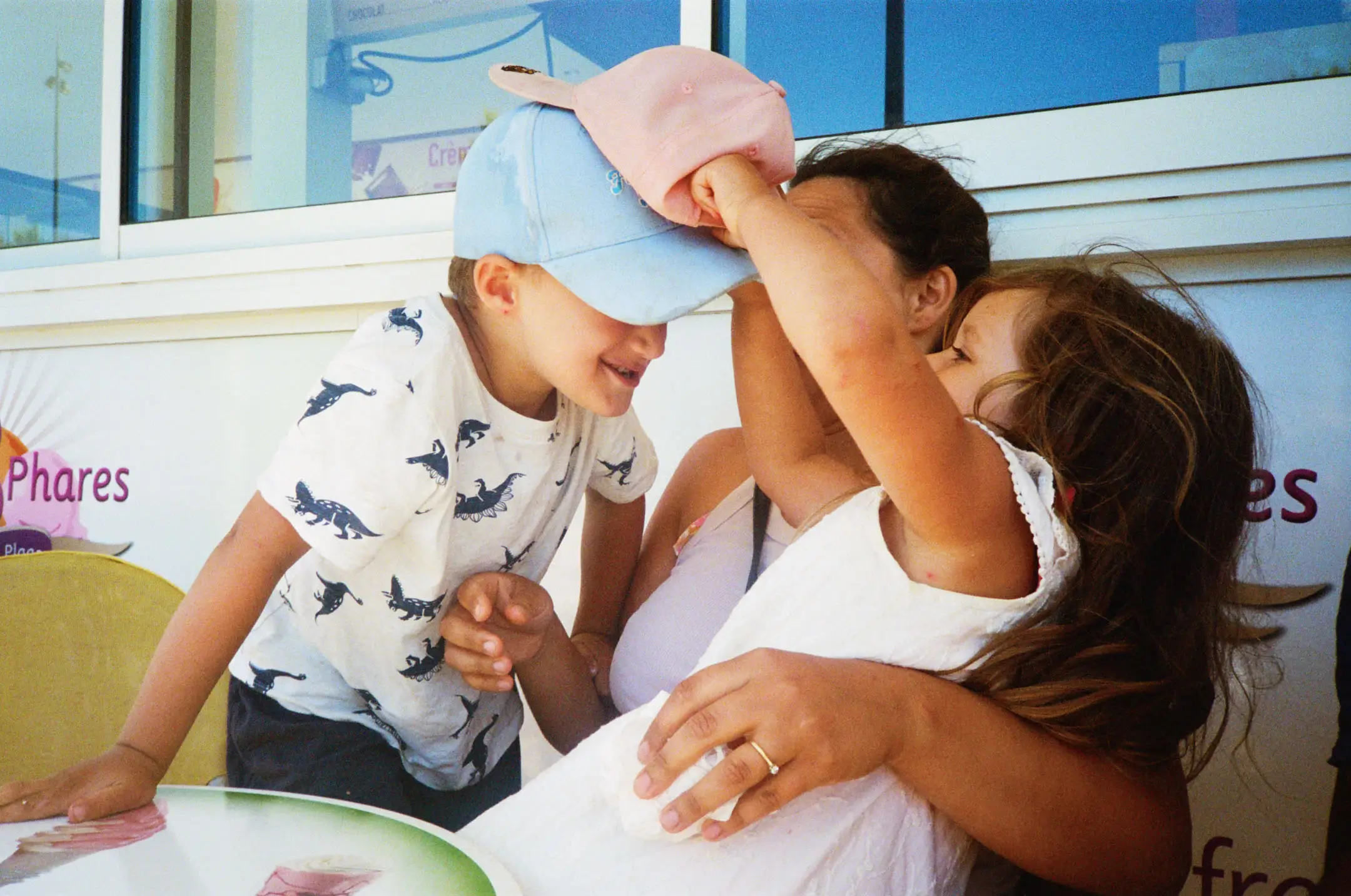 A woman embraces two children as one child playfully puts a pink hat on the boy wearing a blue cap and dinosaur print shirt.
