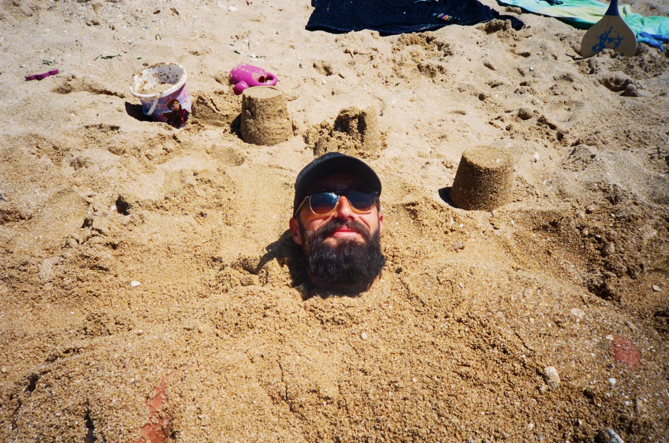 Man buried in sand up to his neck at the beach with three small sandcastles around him and wearing sunglasses and a cap.