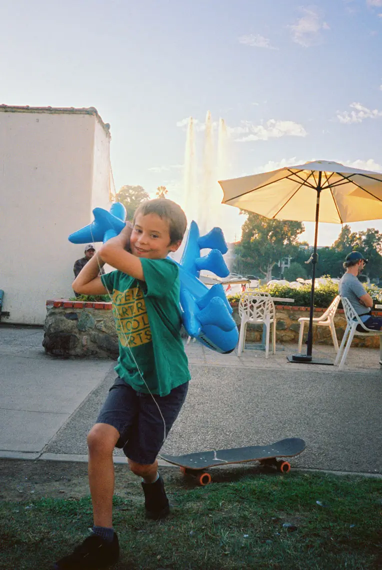 Smiling young boy holding a large blue inflatable airplane while running near a skateboard, with outdoor seating and an umbrella in the background.