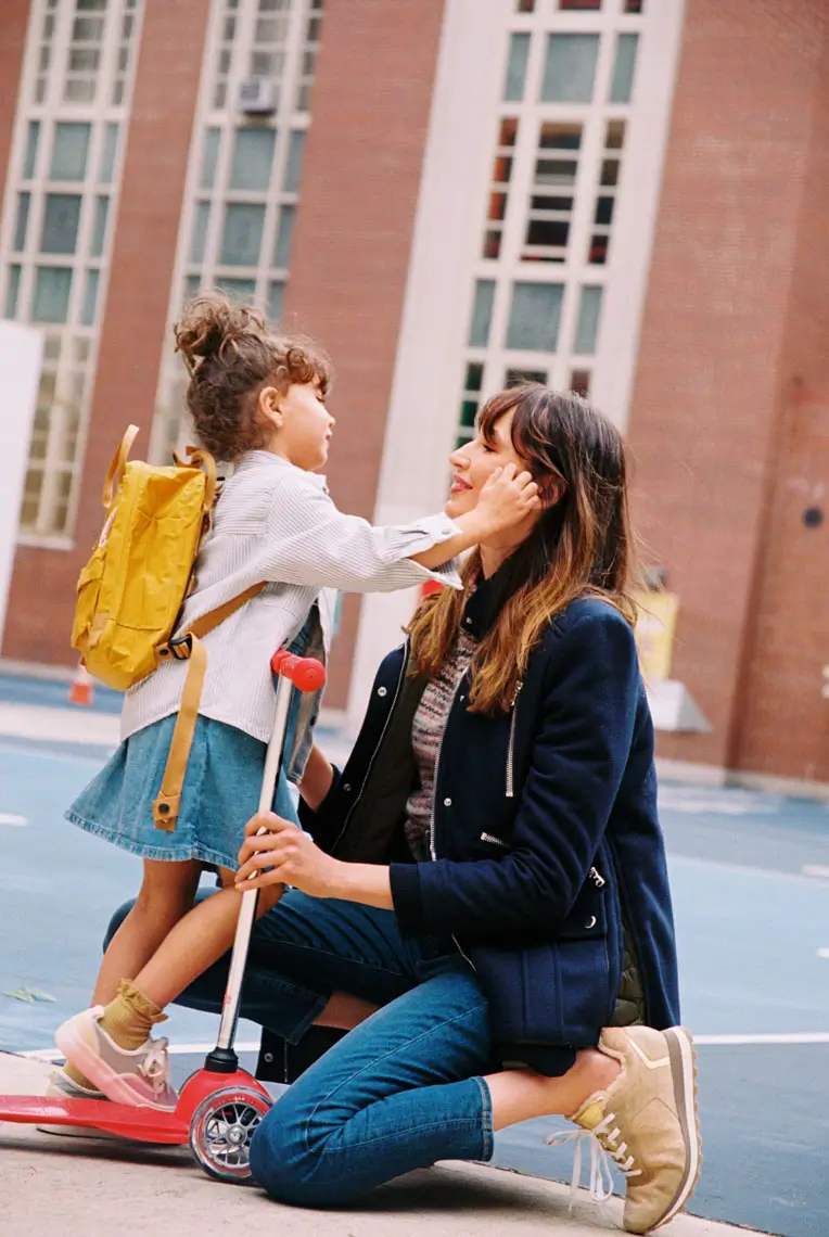 Young girl on a red scooter gently touching a kneeling woman's face in an outdoor urban setting.