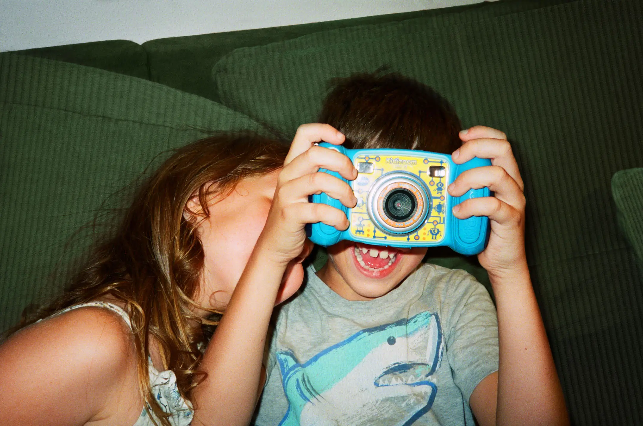Two children sitting on a green couch; a boy holding a blue and yellow toy camera up to his face, smiling widely, and a girl leaning in close to him.