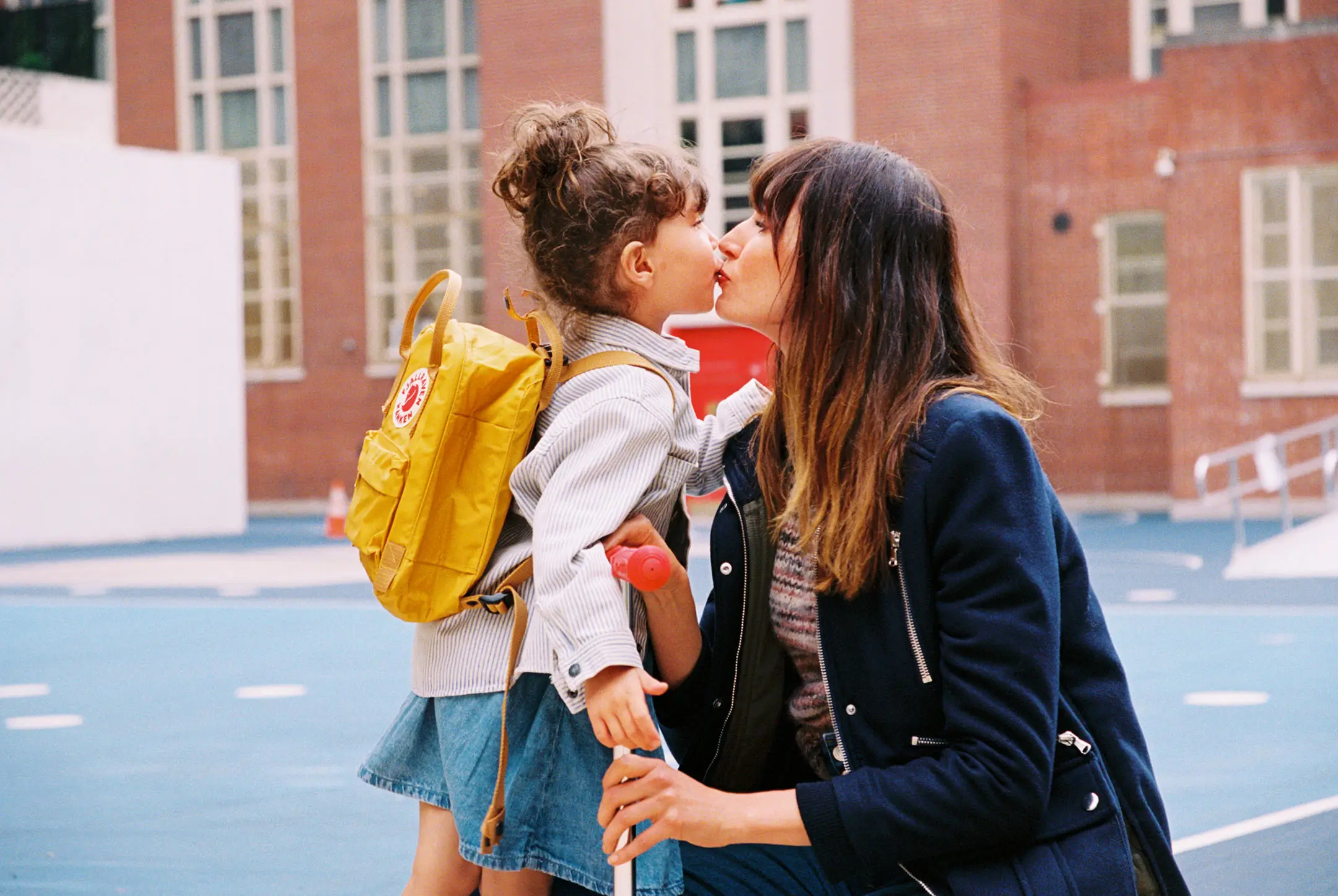 Child with yellow backpack kissing a woman outdoors in front of a brick building.