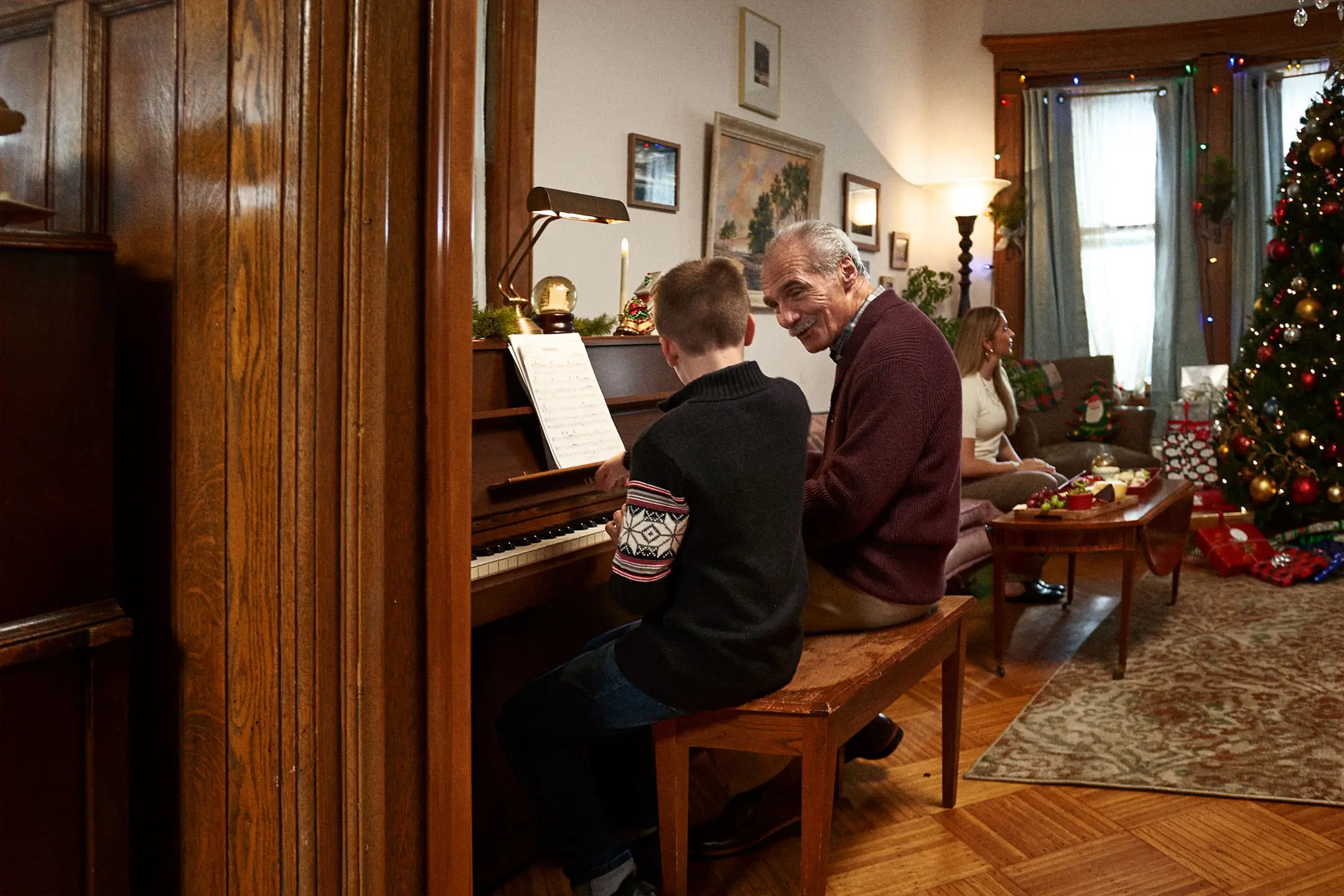An elderly man and a boy playing piano together in a cozy living room decorated for Christmas with a tree and wrapped gifts, while a woman sits nearby.