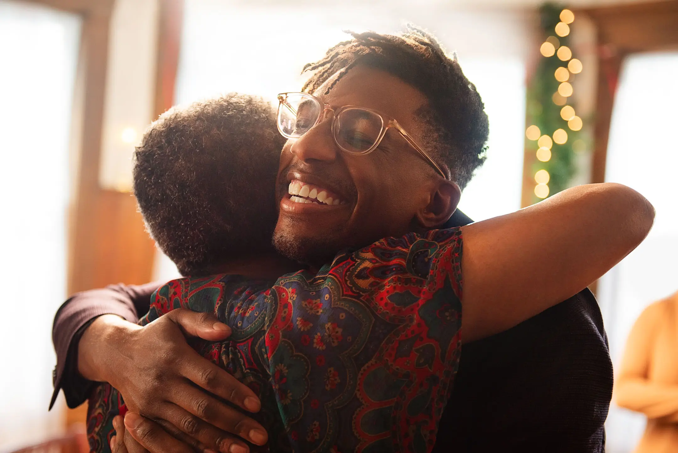 Two people warmly embracing indoors with festive holiday lights in the background.