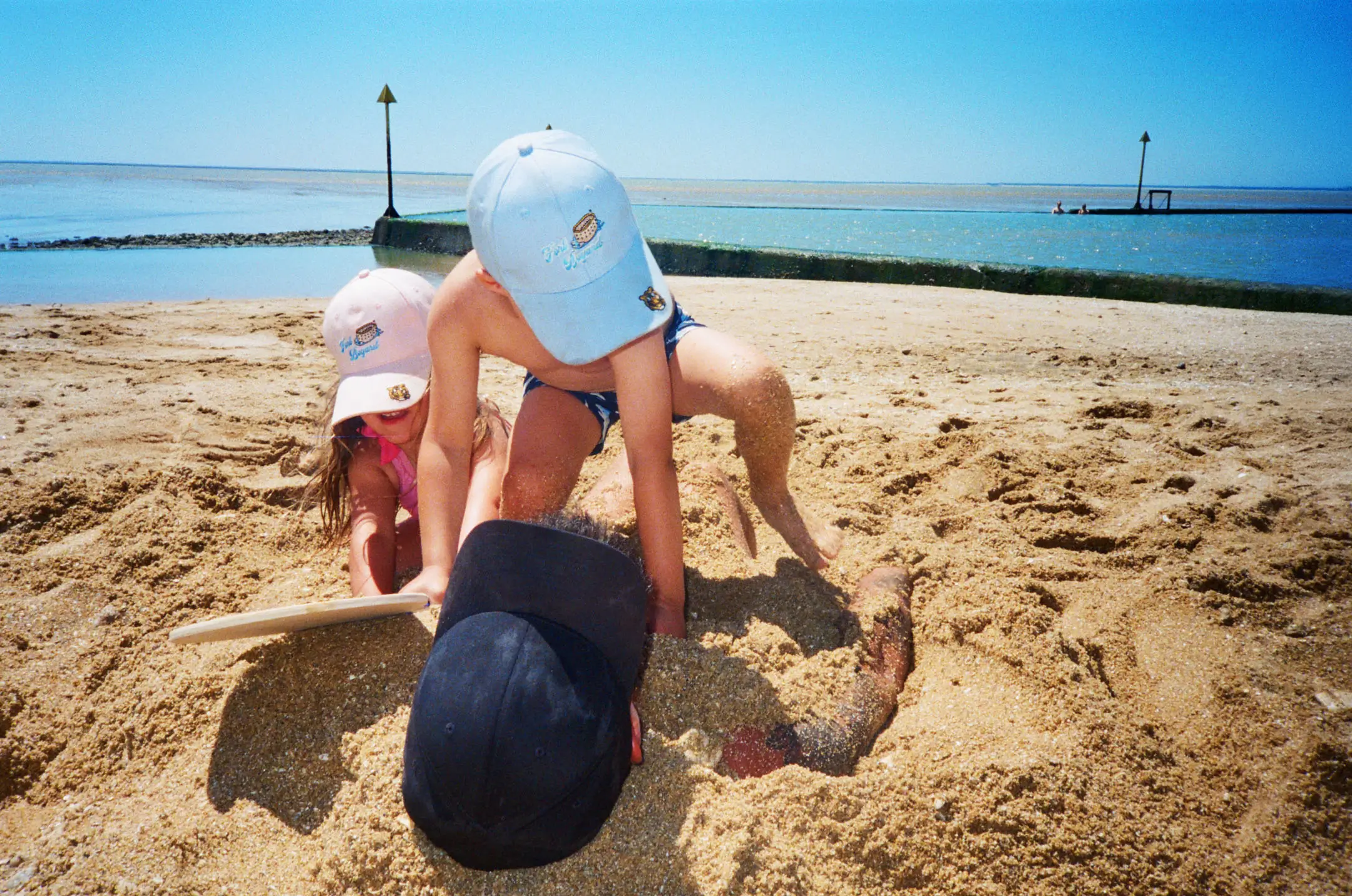Two children wearing hats bury a man in sand on a sunny beach near calm water.