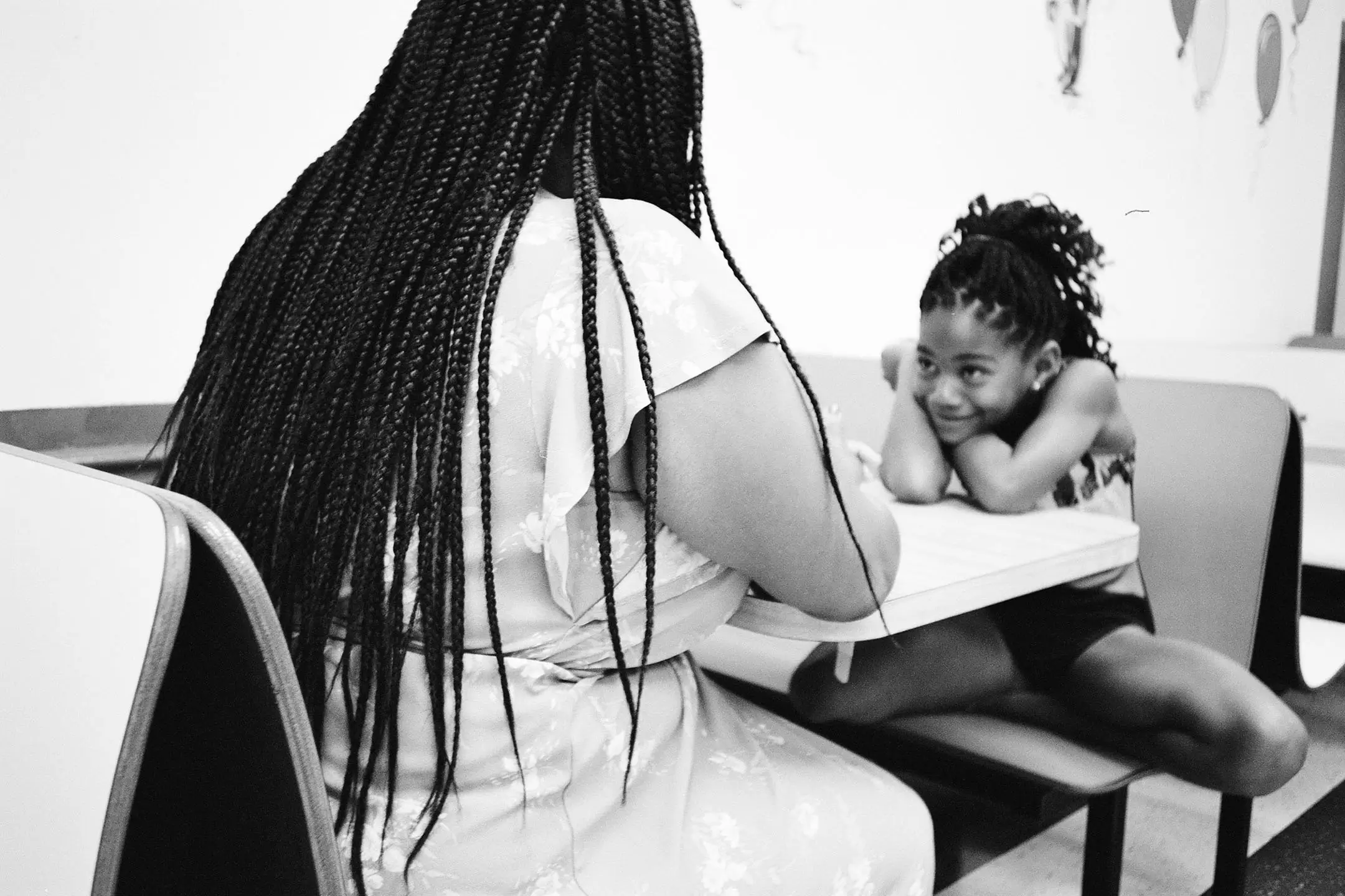 A woman with long braided hair sits across from a smiling young girl leaning on a table in a casual setting.