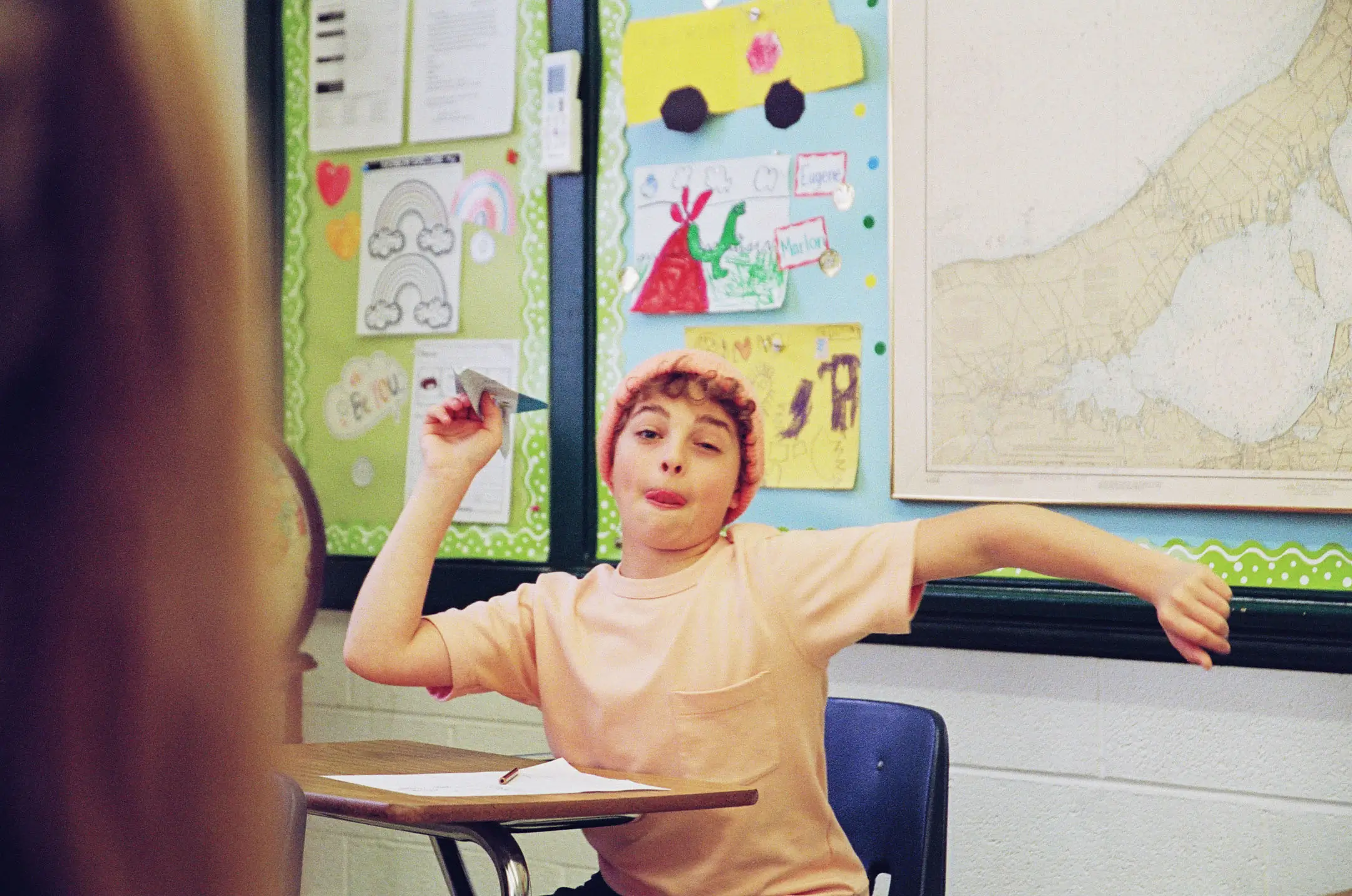 Child wearing a pink beanie and peach shirt throwing a paper airplane in a colorful classroom.