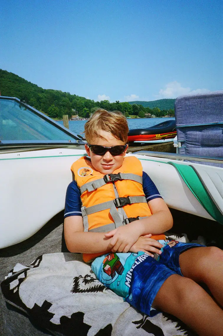 Young boy wearing sunglasses and an orange life jacket, sitting on a boat with a Minecraft-themed towel and lake and forest in the background.