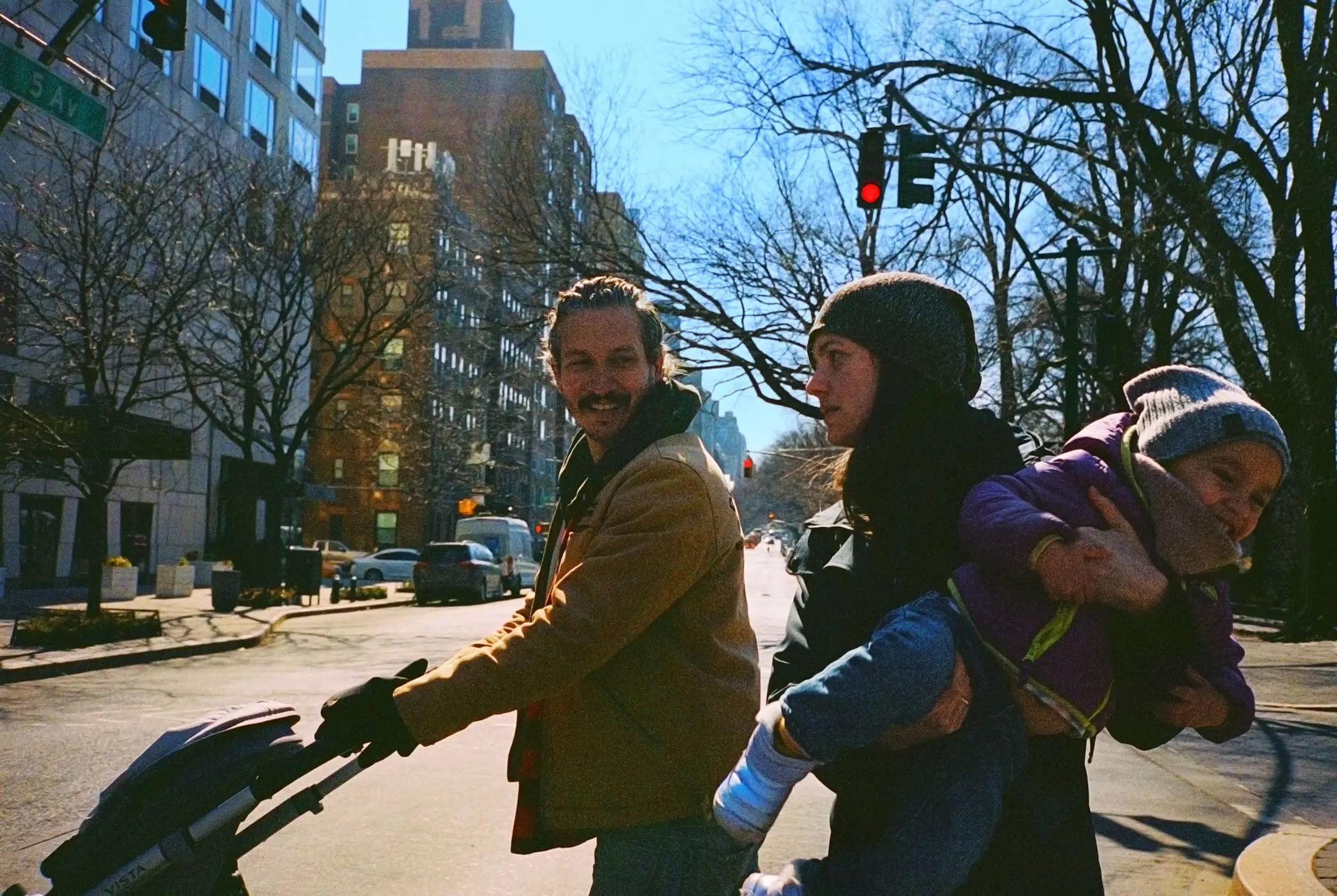 Man pushing a stroller and smiling at a woman holding a laughing toddler on a sunny city street with bare trees and buildings.