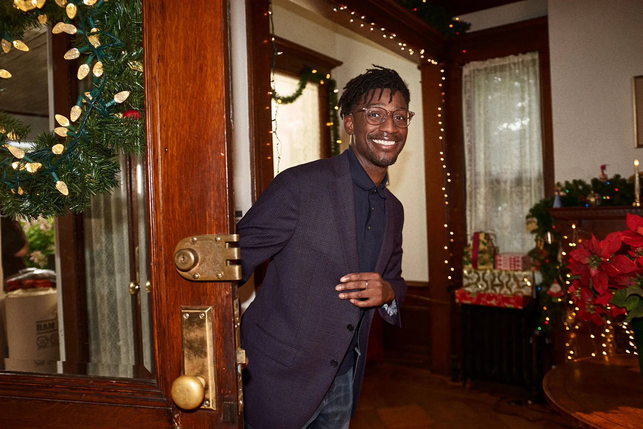Smiling man wearing glasses and a dark blazer opening a decorated wooden door into a warmly lit room with holiday decorations and wrapped gifts.