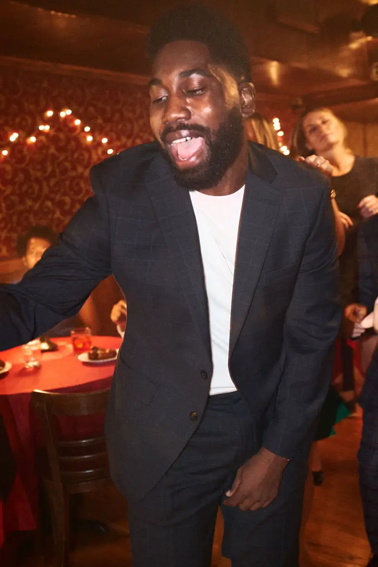 Smiling man in a dark suit jacket dancing joyfully at a festive holiday party with people and decorated tables in the background.