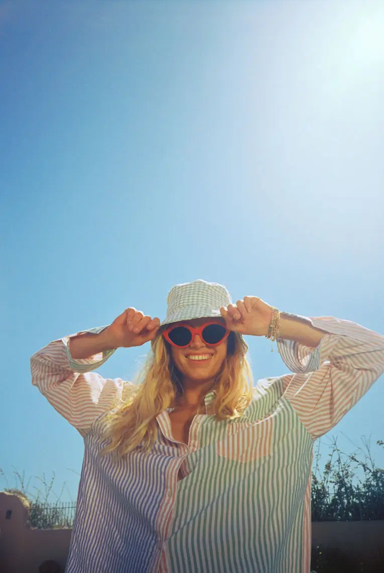 Smiling woman wearing red sunglasses, a checkered bucket hat, and a striped shirt under bright blue sky.