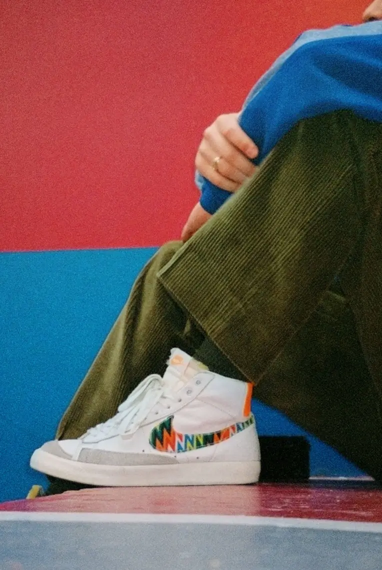 Person wearing white high-top Nike sneakers with colorful zigzag swoosh and olive green corduroy pants against a red and blue background.