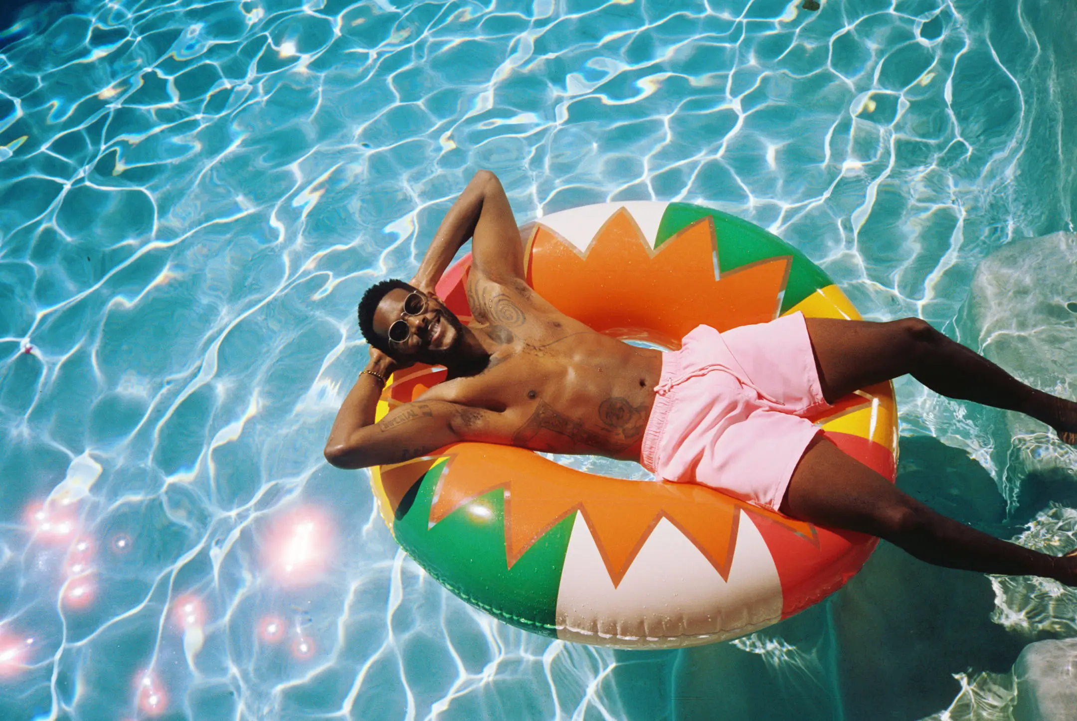 Smiling man wearing sunglasses and pink shorts relaxing on a colorful inflatable ring in a swimming pool.