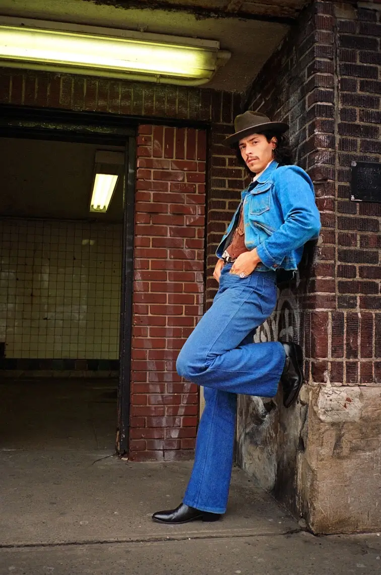 Man wearing a brown hat, denim jacket, and blue jeans leaning against a brick wall with one leg raised.