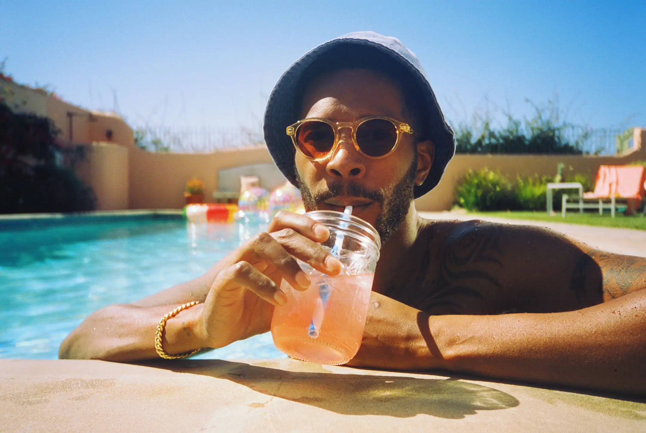 Man wearing round sunglasses and a bucket hat sipping a pink drink from a mason jar by a sunny pool.