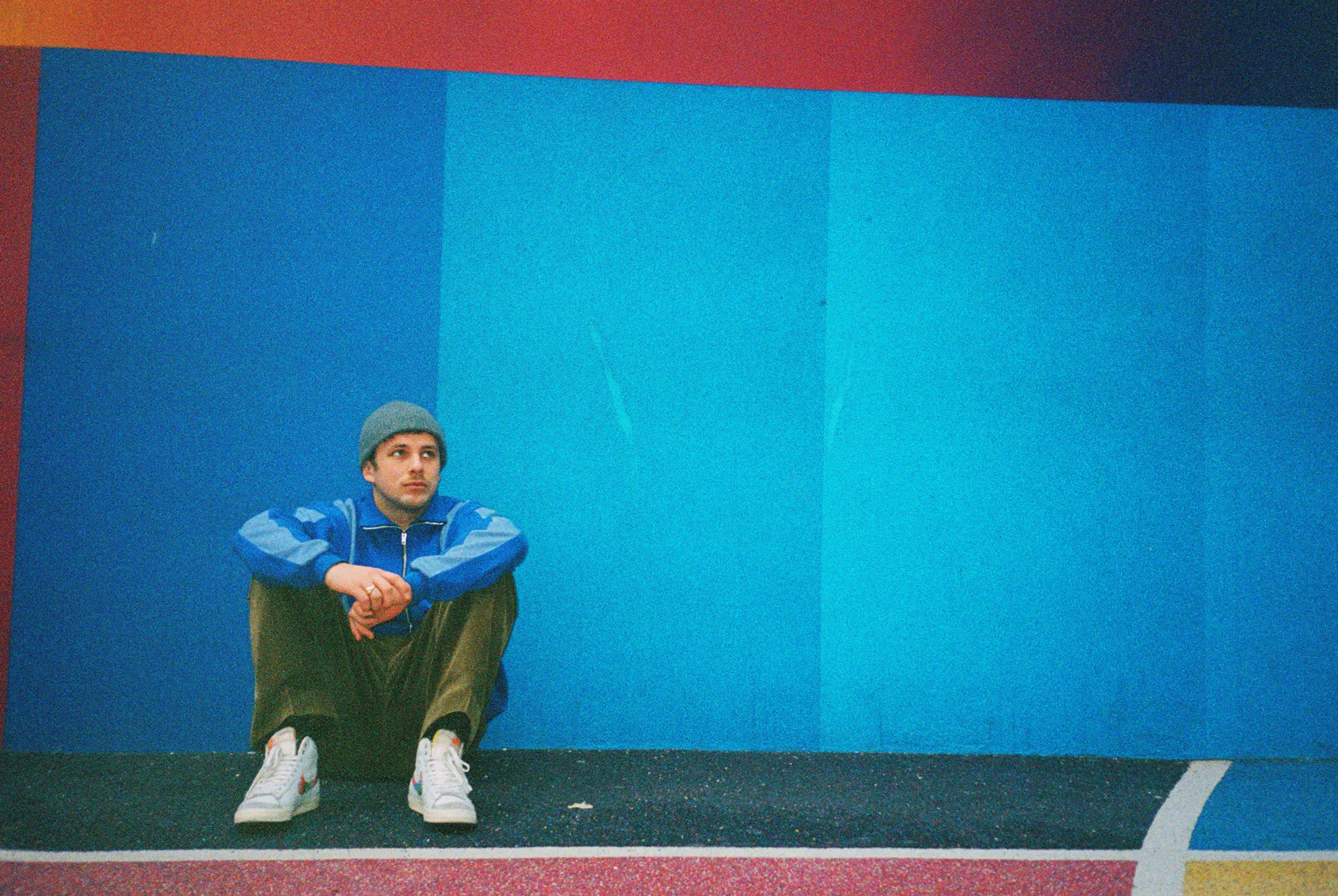 Man in a blue jacket and gray beanie sitting on the ground against a vibrant blue and red geometric wall.