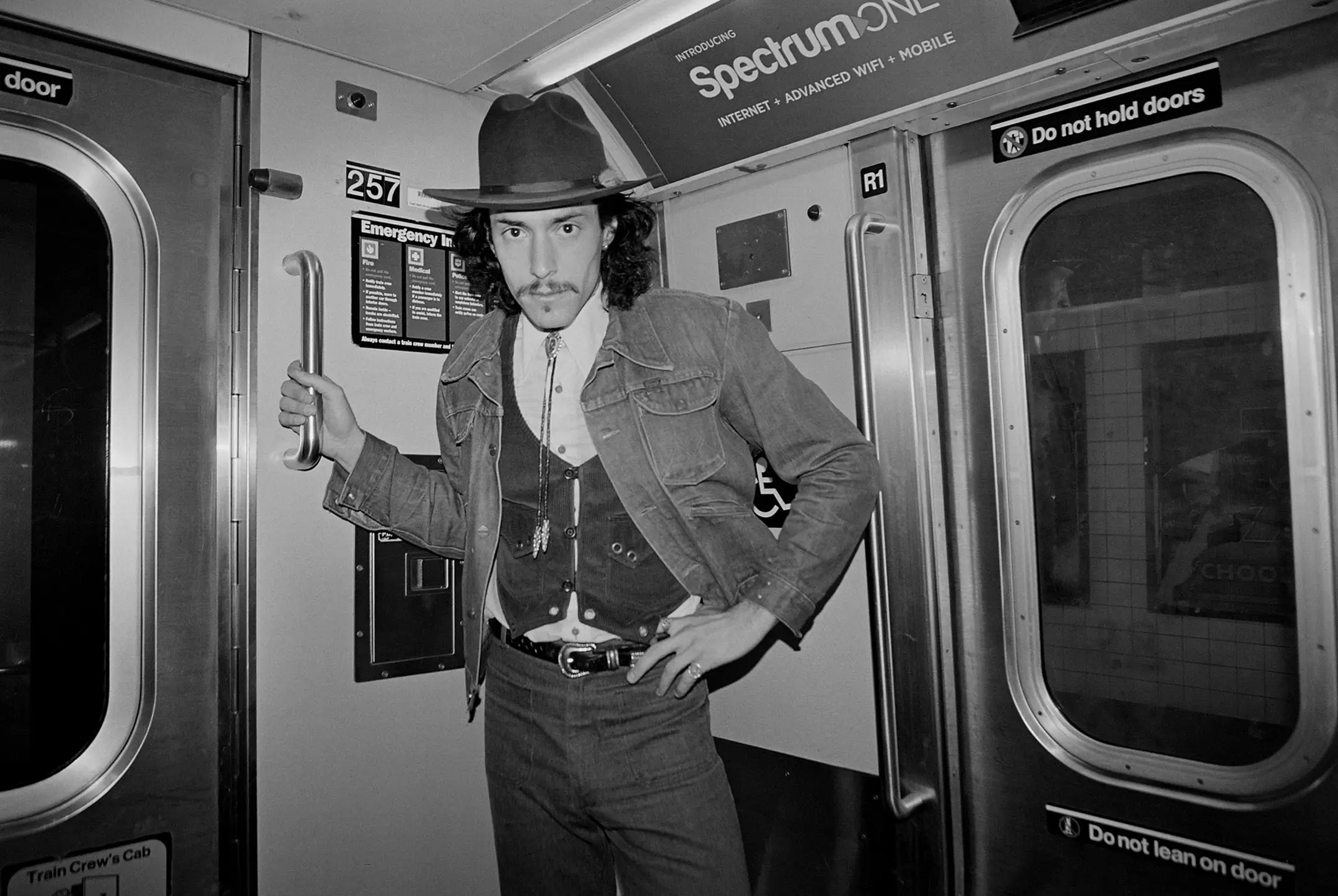 Man wearing a fedora hat, denim jacket, and vest standing inside a subway train holding a metal handle.