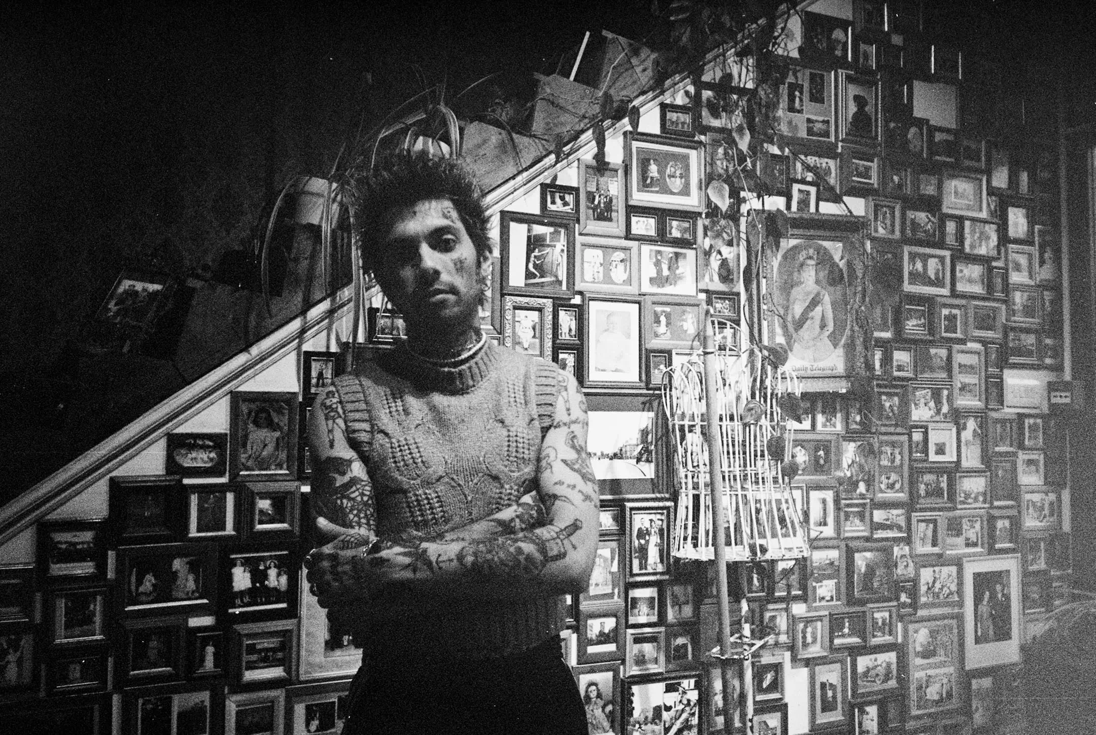 Black and white photo of a tattooed person with spiked hair and a sleeveless knit top standing with arms crossed in front of a wall filled with framed photographs and a dress form covered in vines.