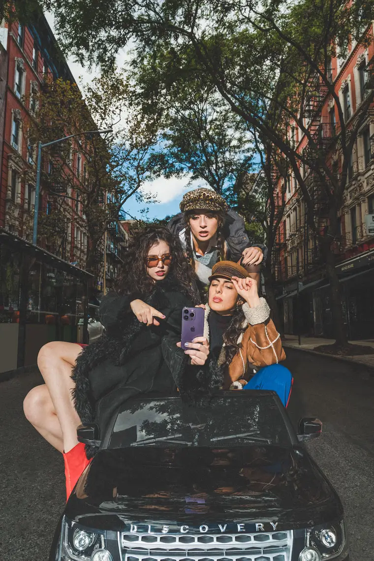 Three stylish young women posing for a selfie while sitting and standing on a black Discovery car in a city street lined with trees and buildings.