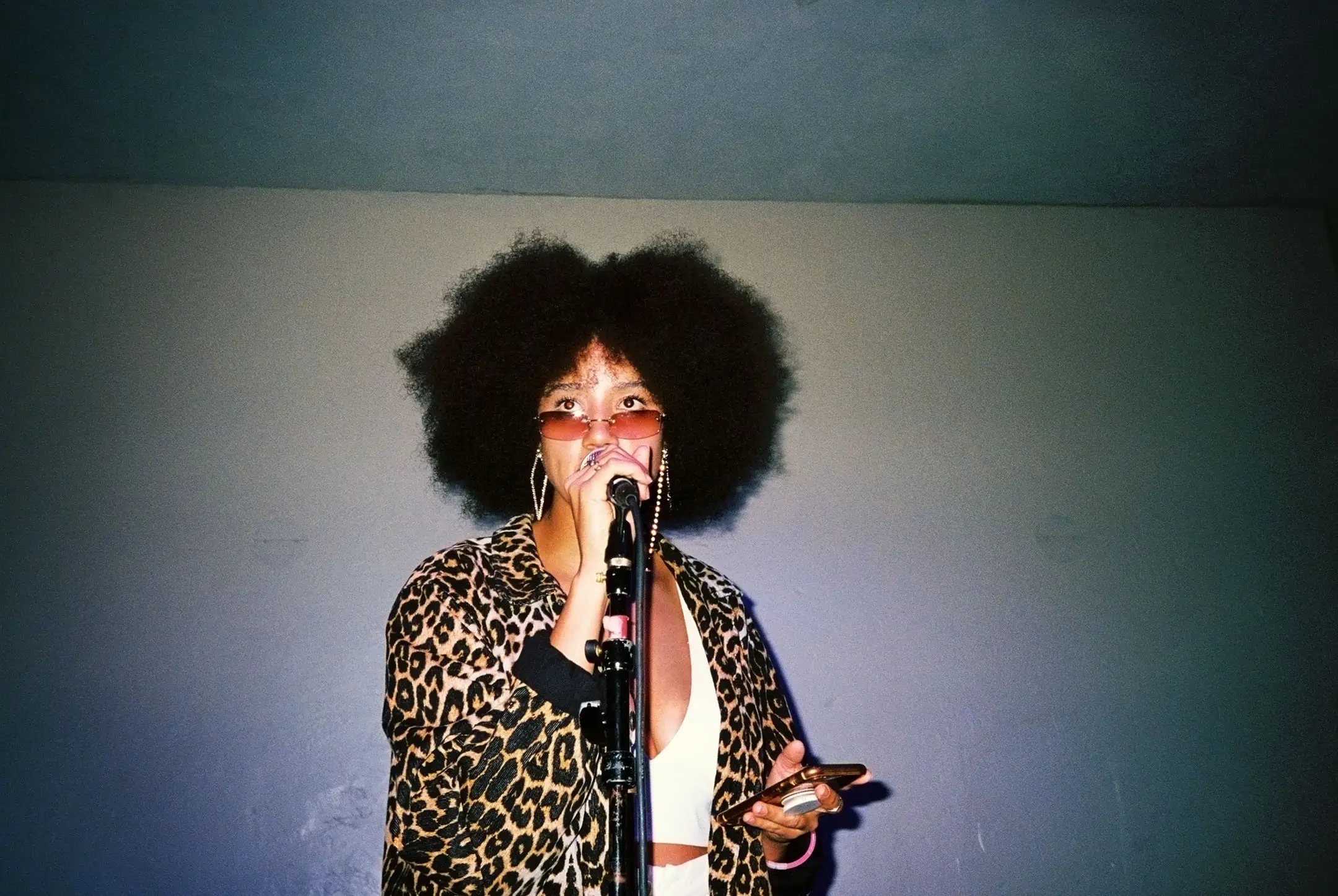 Woman with large afro wearing leopard-print jacket and sunglasses, singing into a microphone while holding a phone.