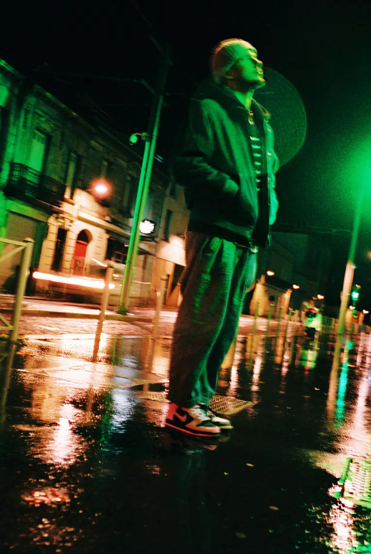 Person wearing a beanie, jacket, and sneakers standing on a wet street at night with green streetlights reflecting on the pavement.