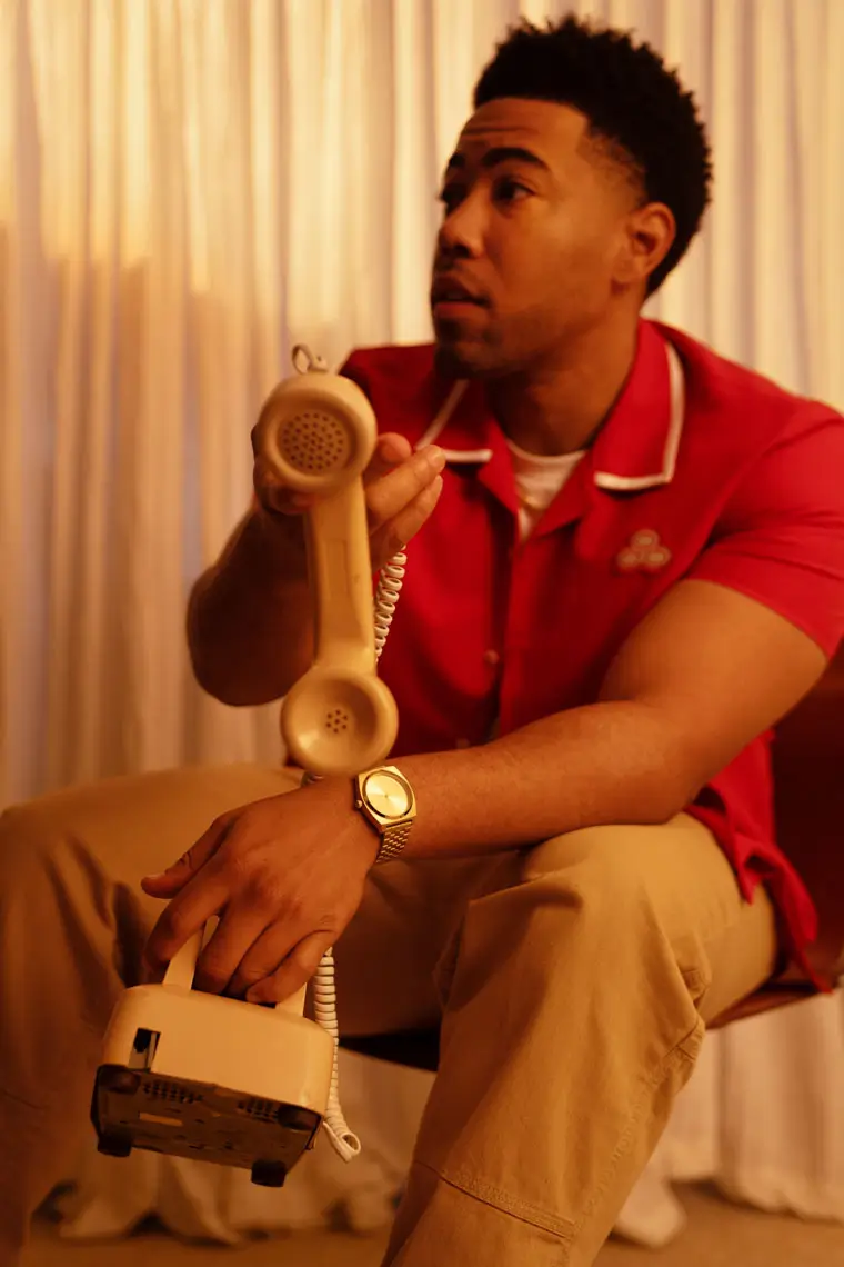 Man in red shirt and tan pants sitting while holding up the receiver of a beige rotary phone.